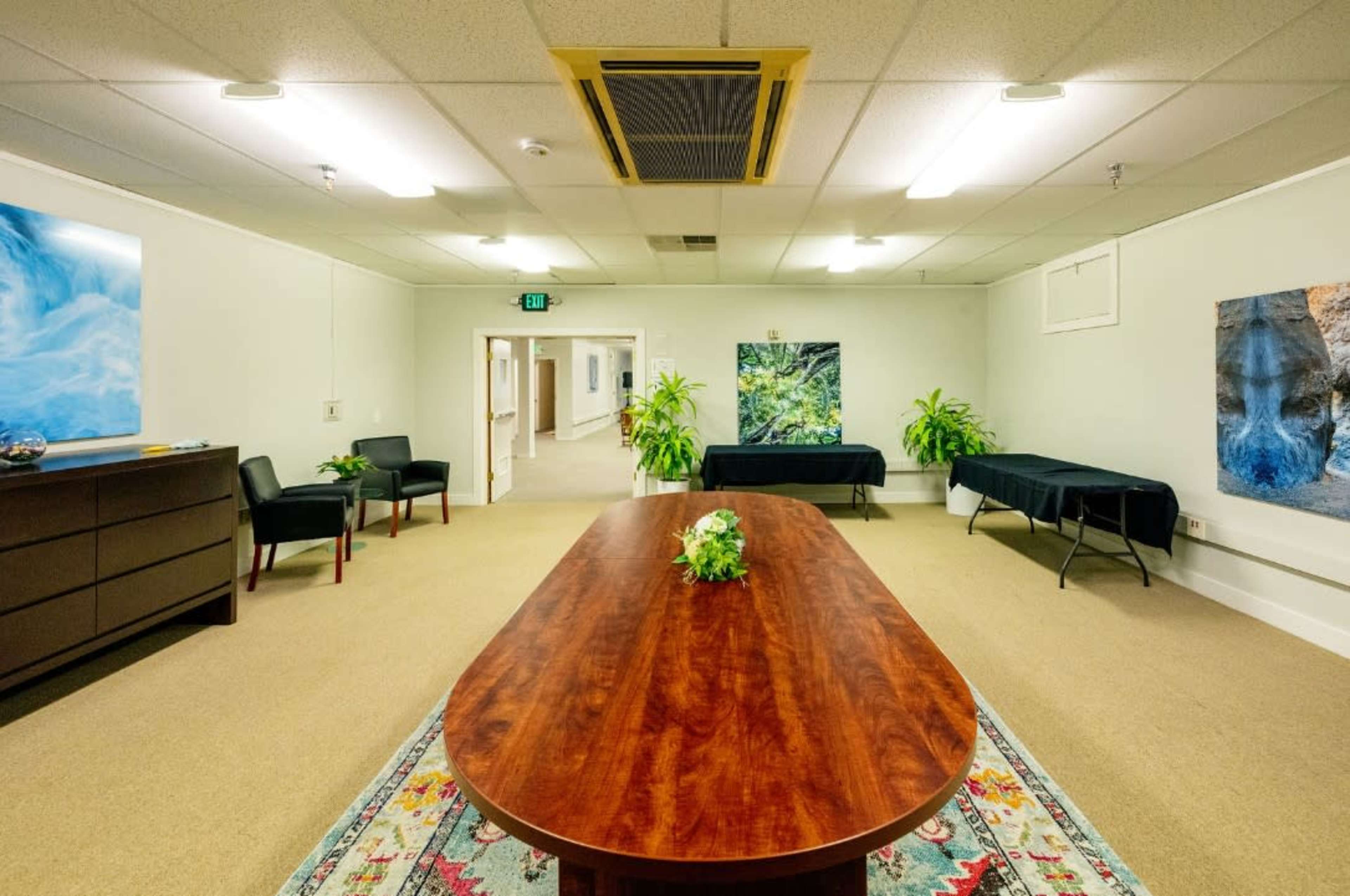 A spacious meeting room features a large wooden table surrounded by chairs, with two plants and a few decorative artworks on the walls.