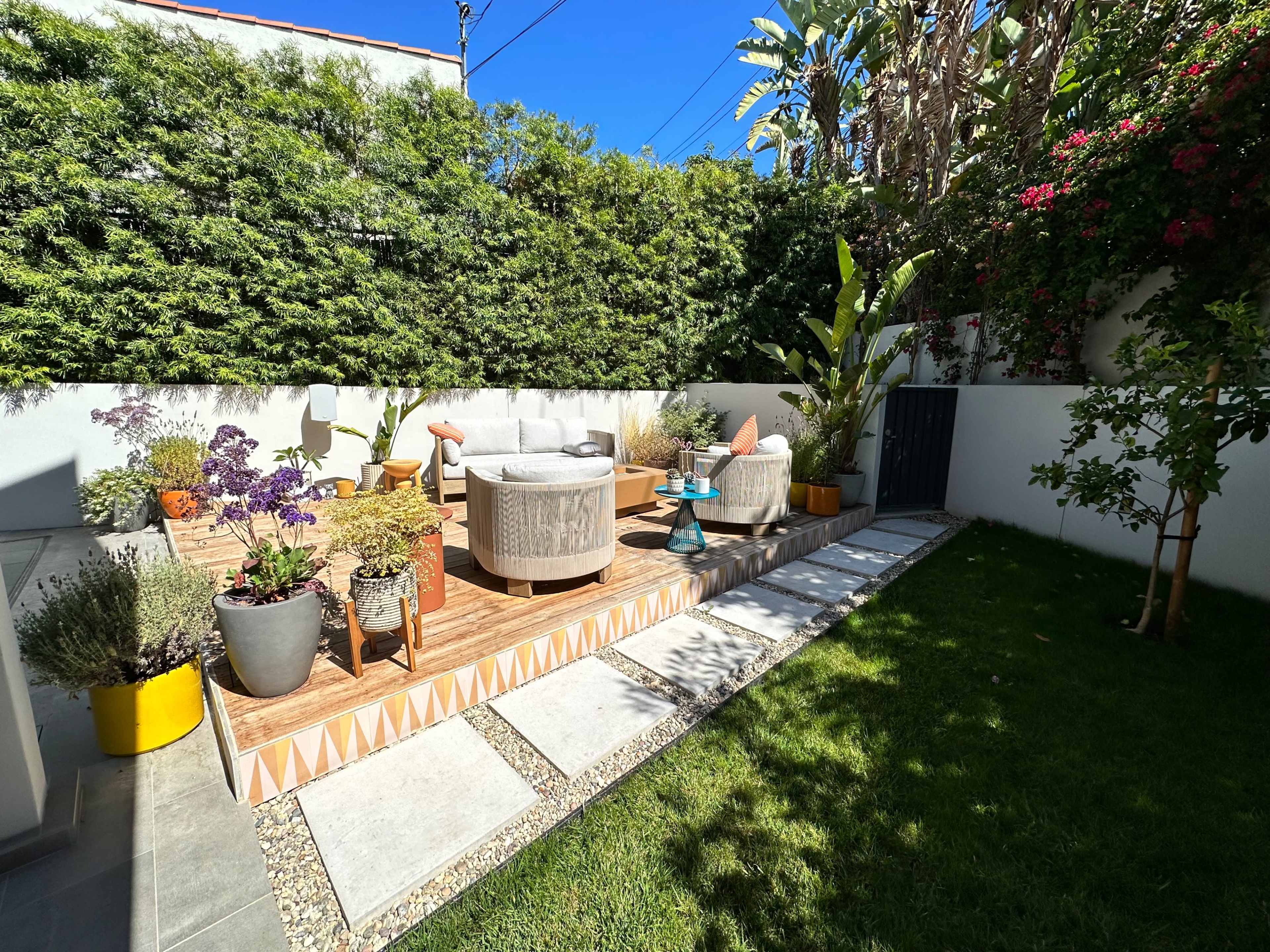 The image shows a landscaped backyard with circular seating arrangements on a wooden deck, surrounded by potted plants and a green lawn.