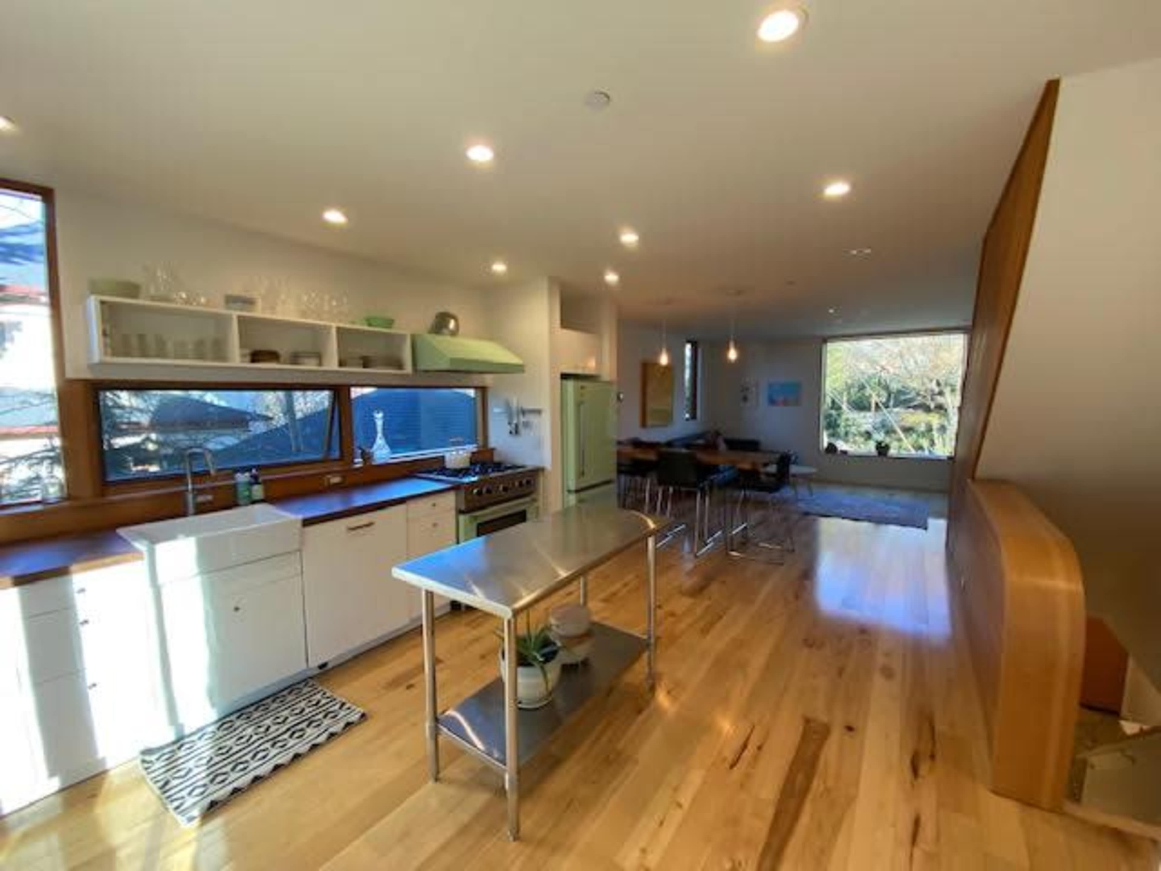 A modern kitchen with wooden flooring, featuring an island, stainless steel appliances, and large windows allowing natural light to enter.
