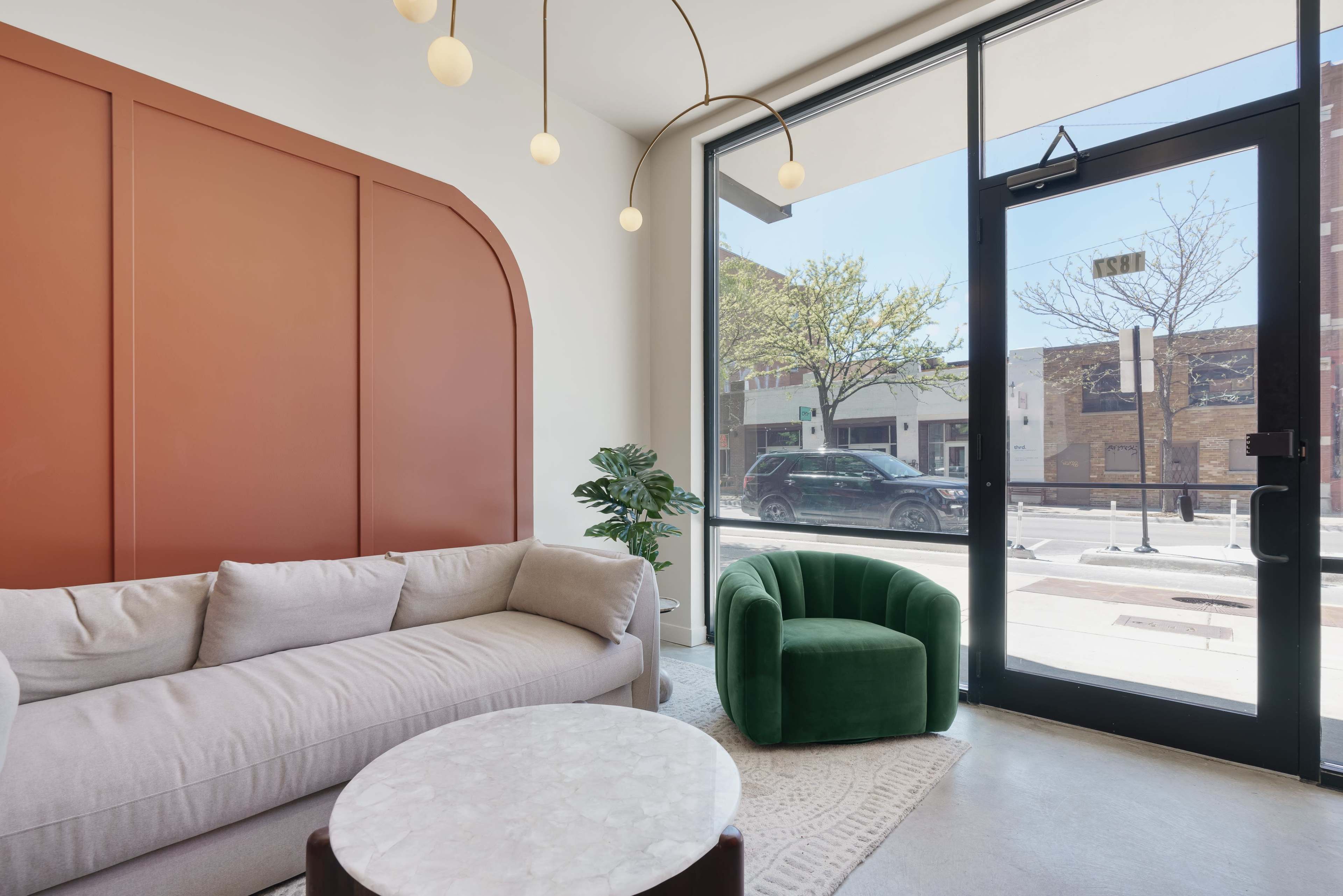 A modern interior space featuring a light-colored sofa, a green armchair, a round marble coffee table, and a large window with street views.