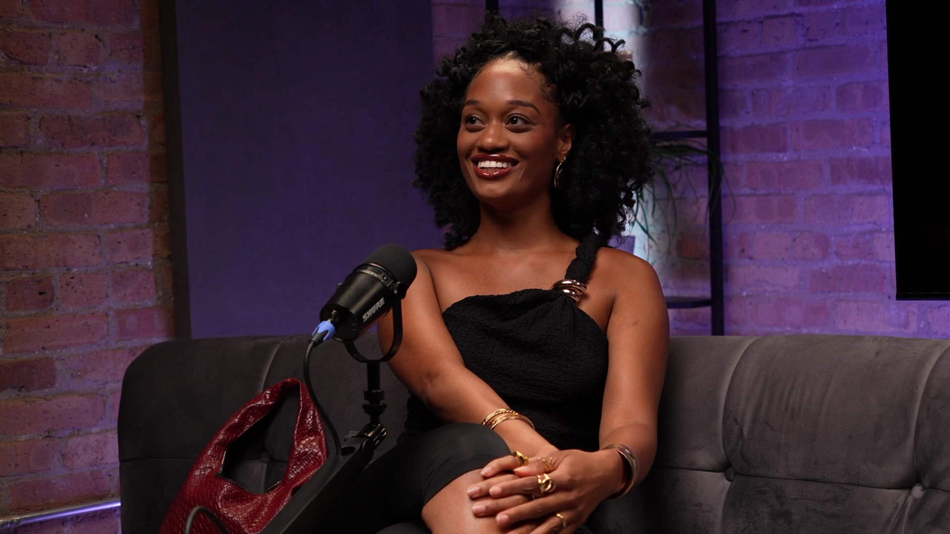 A woman with curly hair sits on a couch next to a microphone, smiling during a podcast recording.