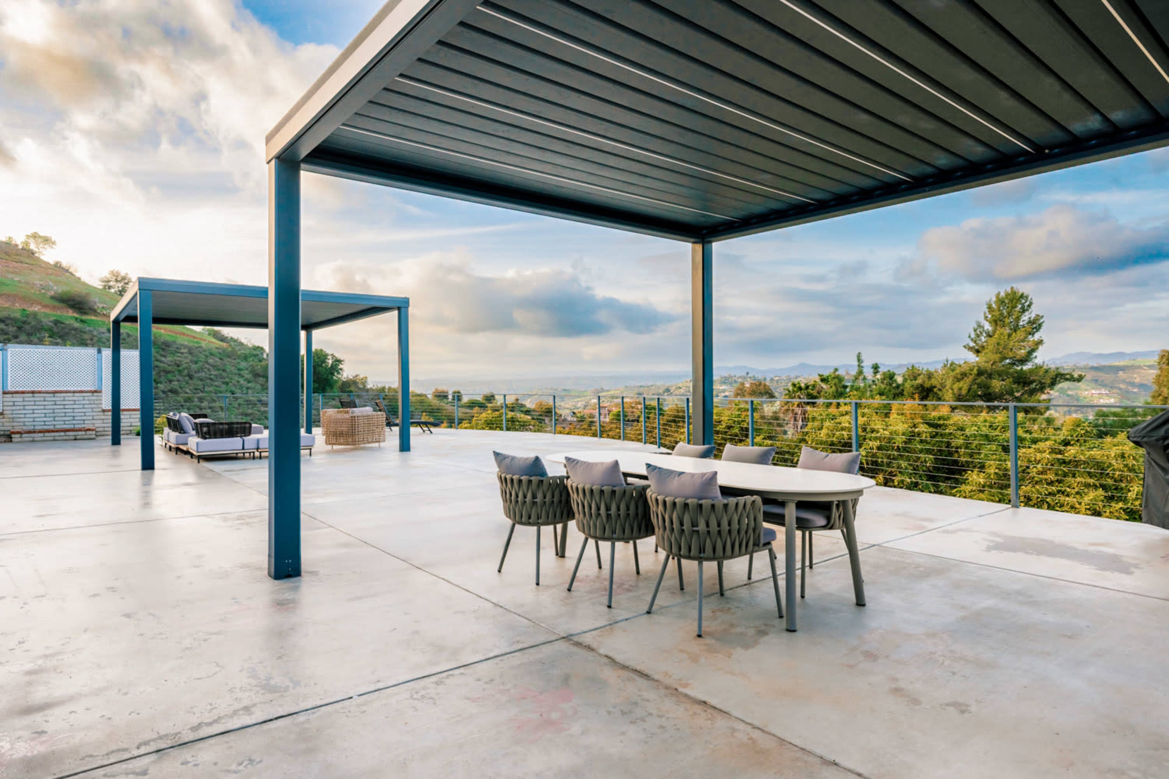 A spacious outdoor patio features a dining table surrounded by chairs, with a view of hills and clouds in the background.