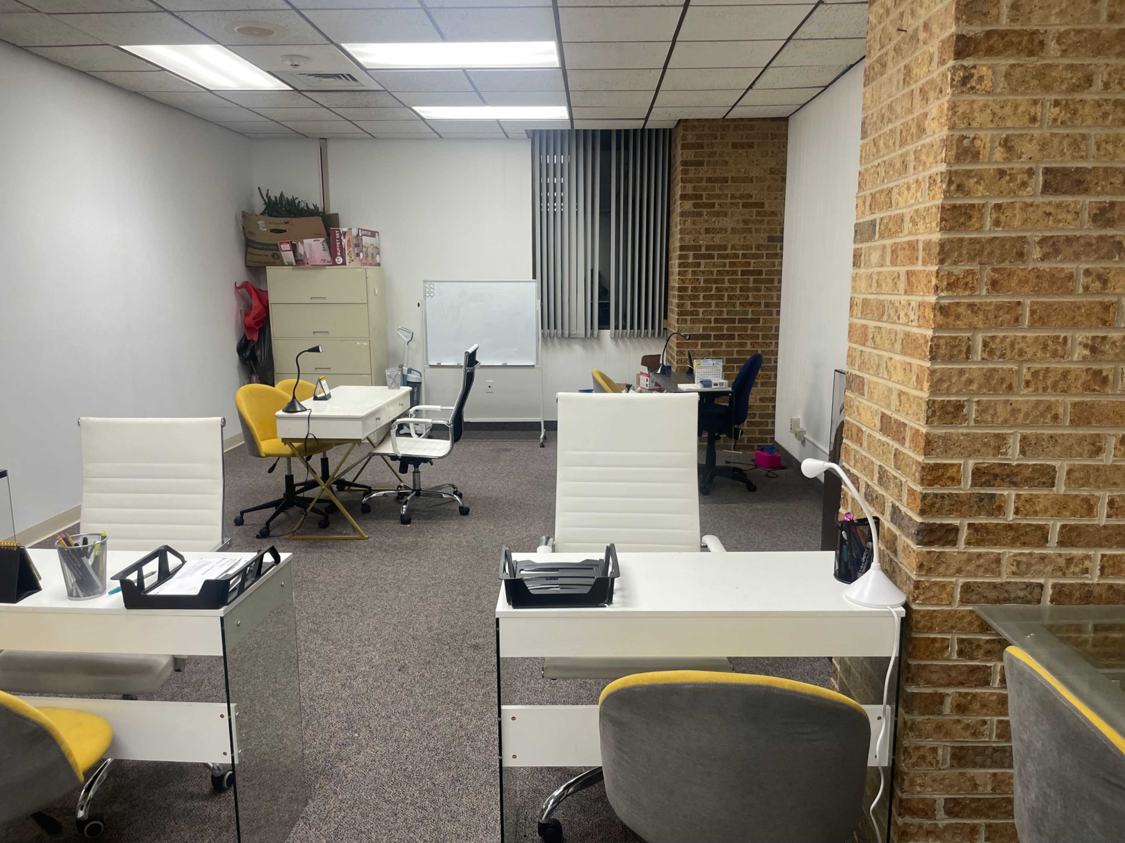 A spacious office area with multiple desks, some equipped with white chairs and others with gray ones, and a brick wall on one side.