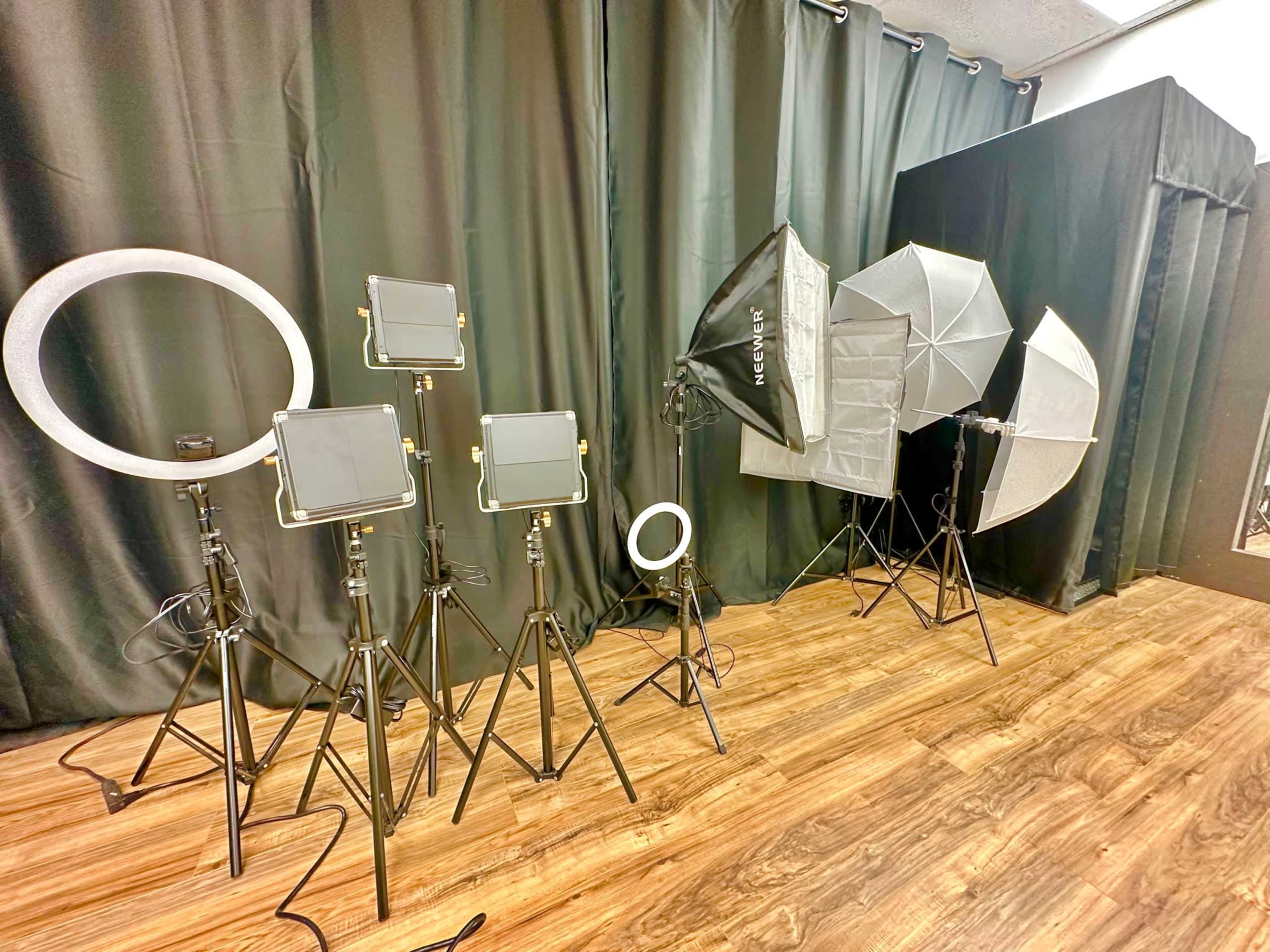 A variety of lighting equipment, including softboxes and LED panels, is set up against a dark curtain backdrop on a wooden floor.