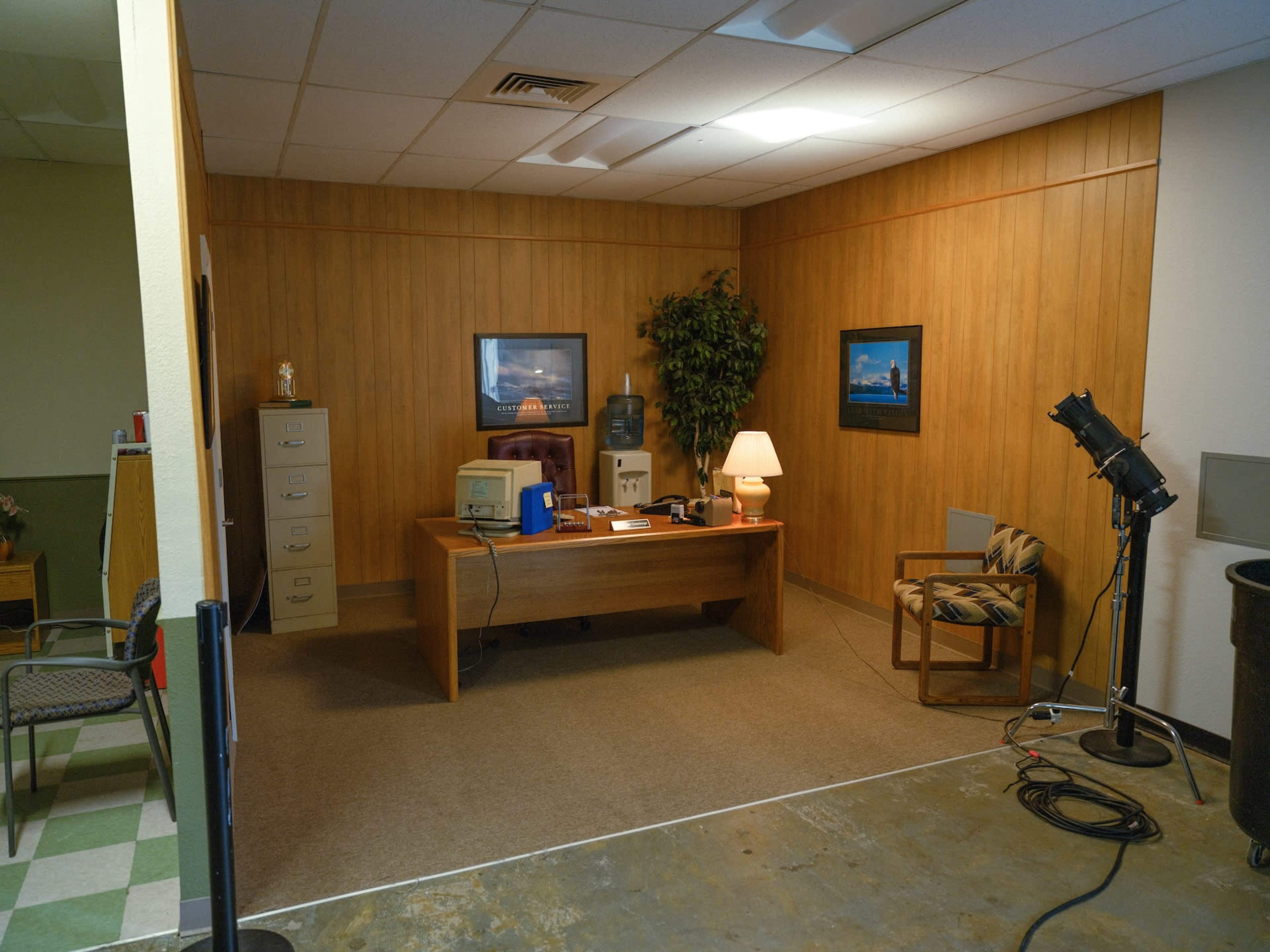 The image shows a simple office space with a wooden desk, filing cabinets, a water cooler, and a chair arranged in a corner with a potted plant and framed pictures on the wall.