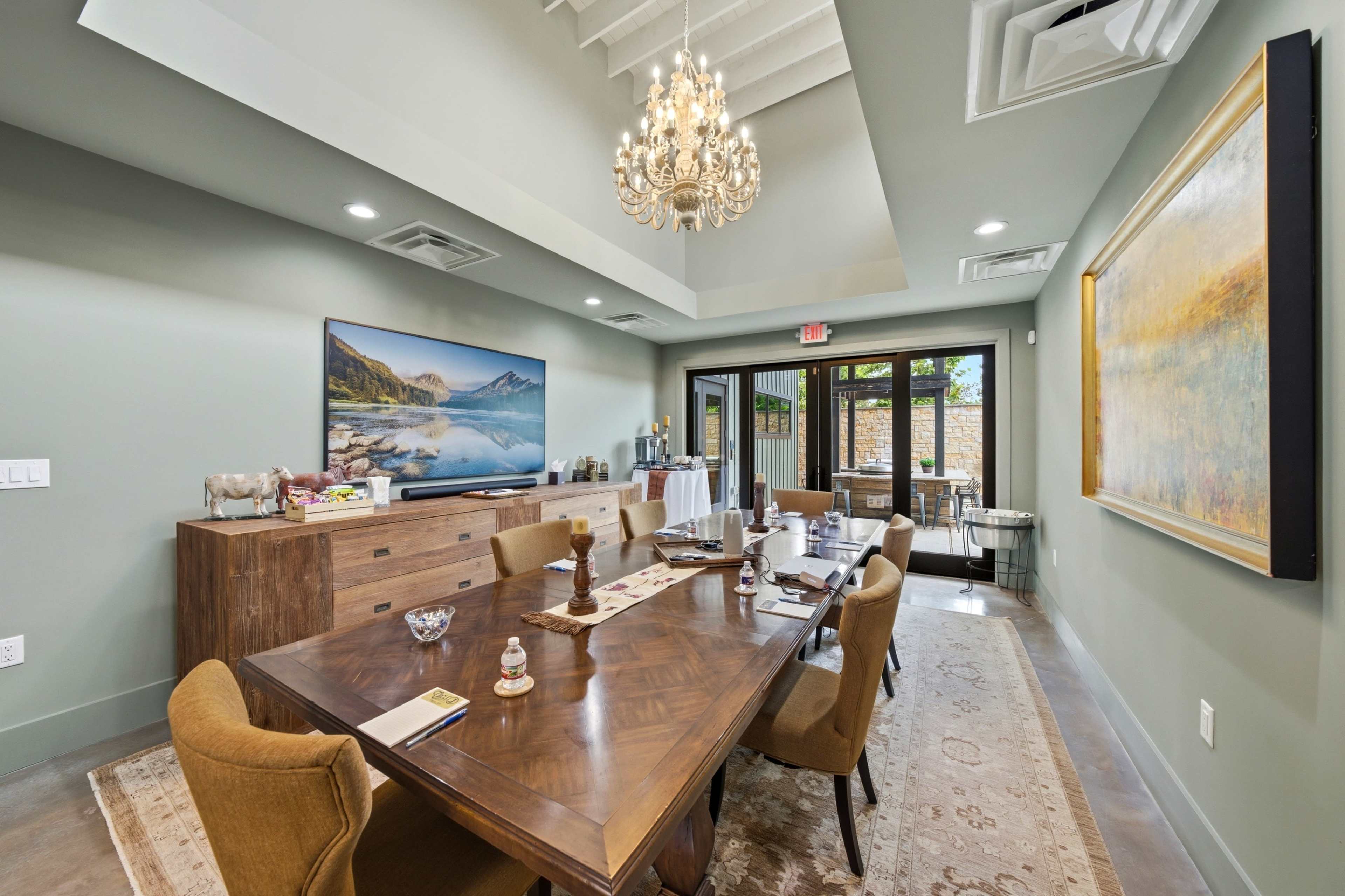 The image shows a modern dining room with a large wooden table set with chairs, a chandelier overhead, and a wall-mounted TV with a landscape picture.