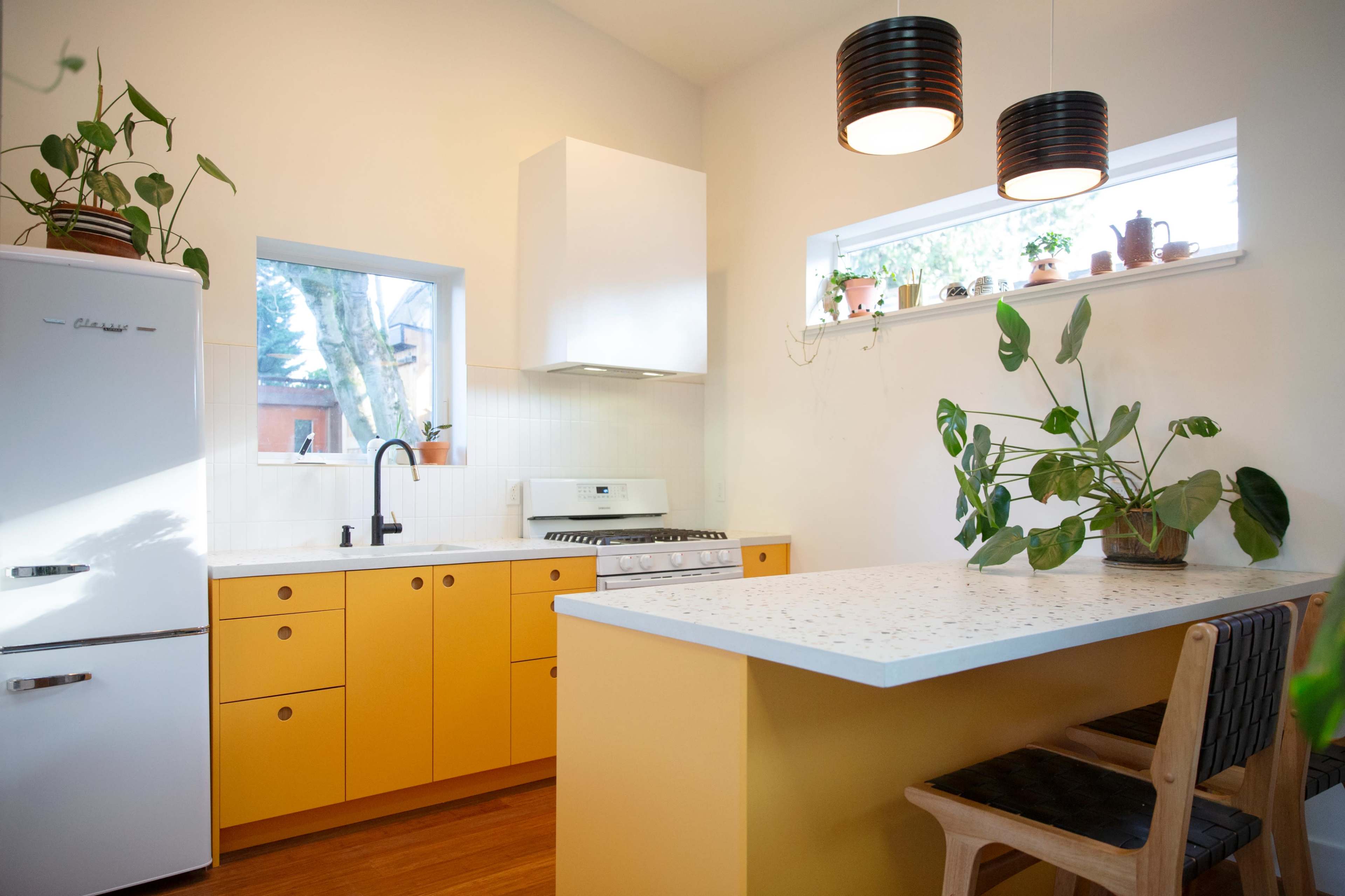 A bright kitchen features yellow cabinets, a white countertop, and a retro refrigerator, with plants decorating the windowsills and countertop.