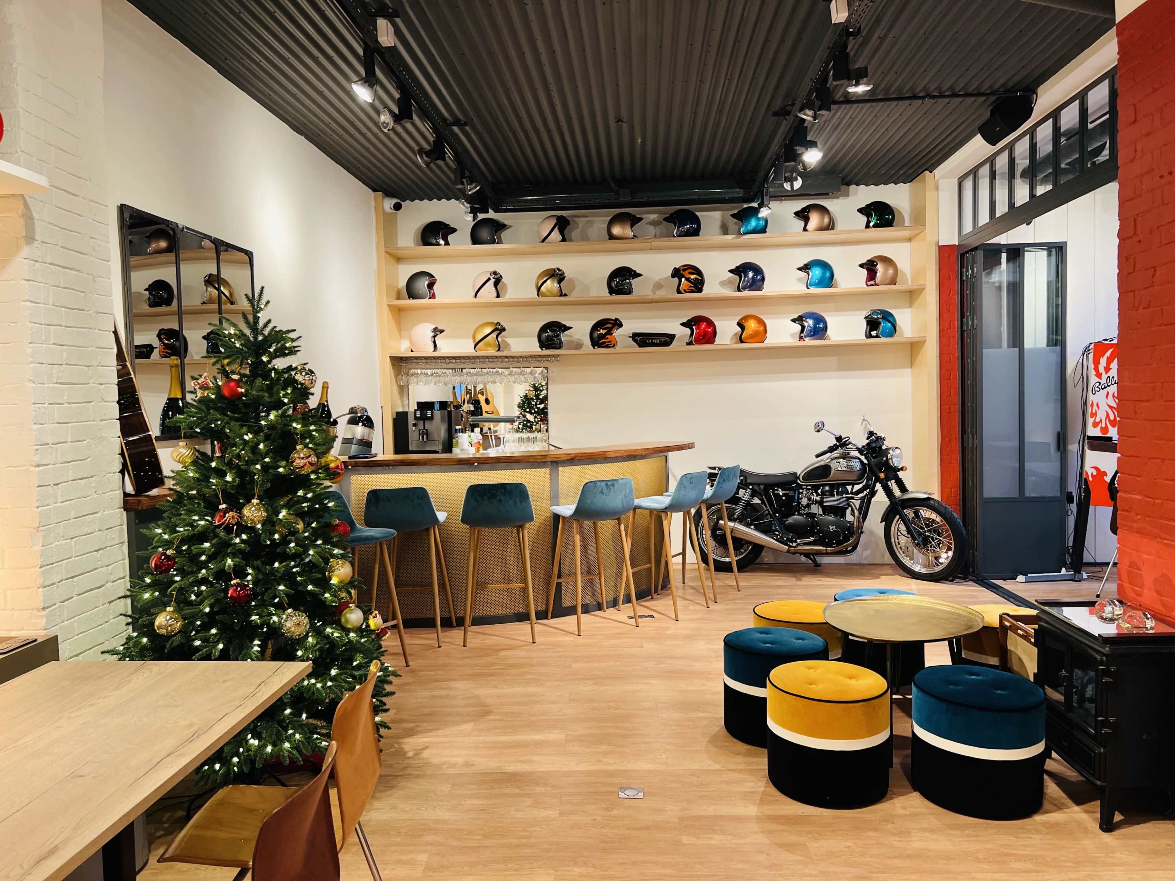 The image shows a stylish interior space with a bar, colorful stools, a Christmas tree, and a motorcycle displayed against a wall of helmets.