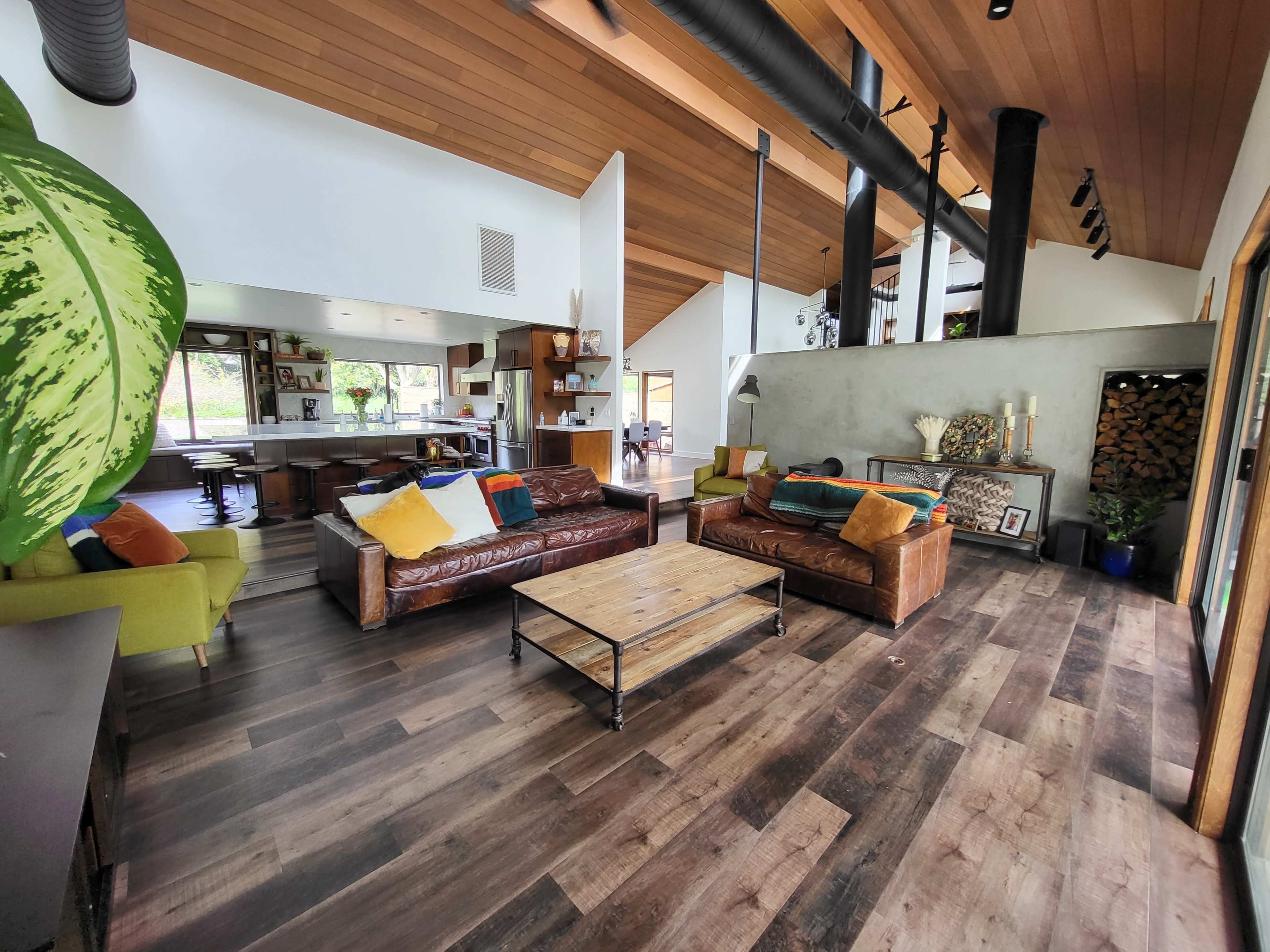 A spacious living room with wooden floors, large windows, and a modern design featuring a mix of leather and fabric sofas, a wooden coffee table, and a view of the kitchen area.
