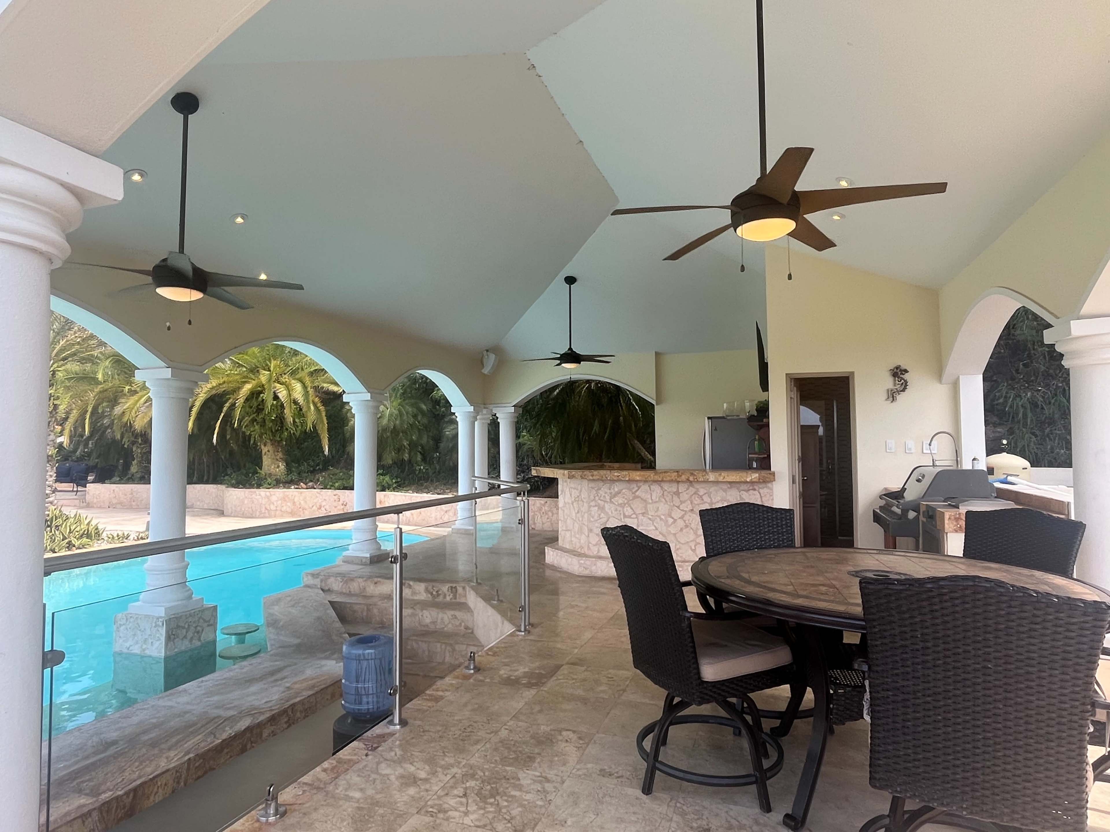 The image shows a covered outdoor patio area with a dining table, two ceiling fans, and a view of a swimming pool surrounded by tropical plants.