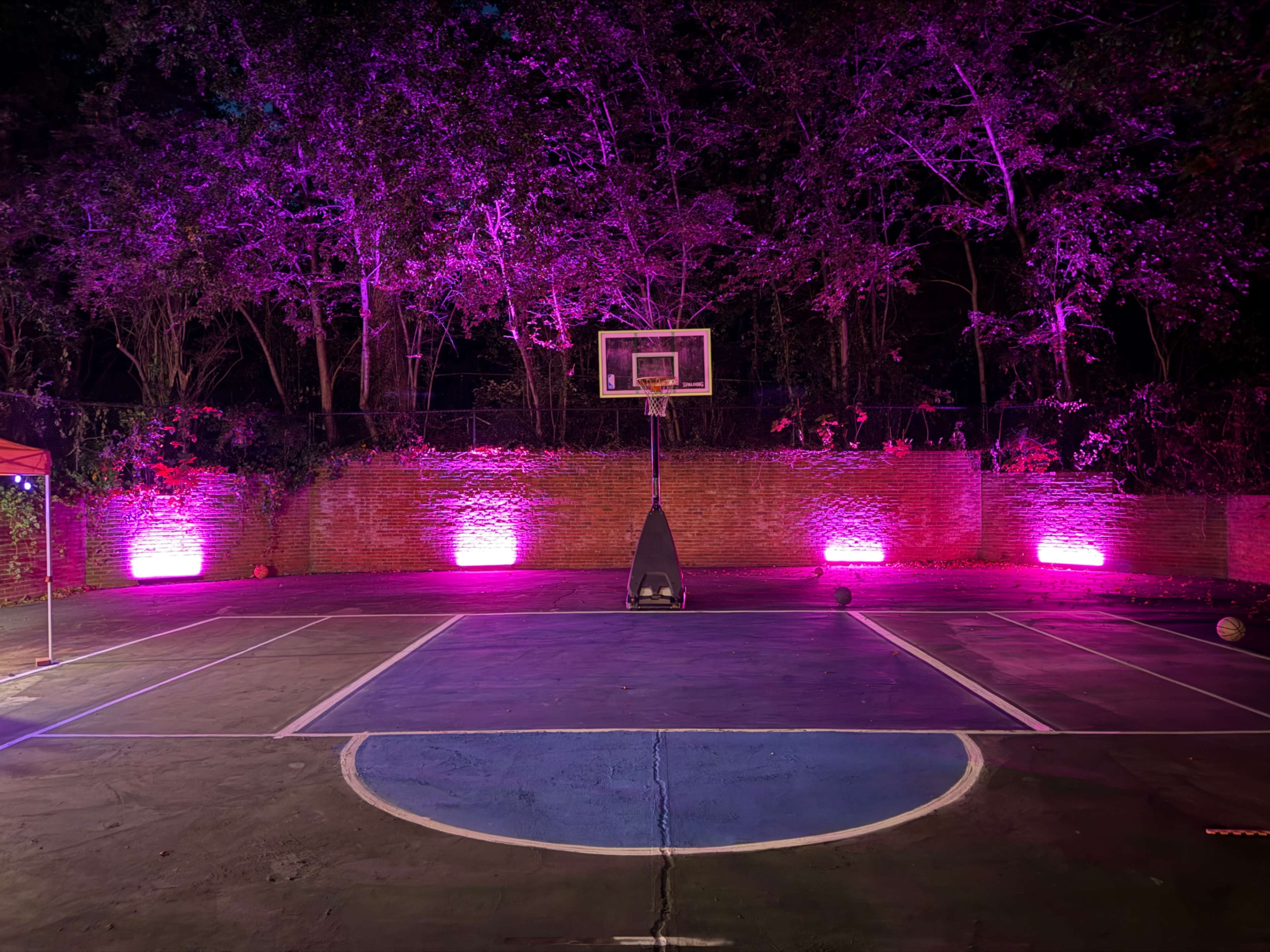 A basketball court is illuminated with purple lights, surrounded by trees and a brick wall.