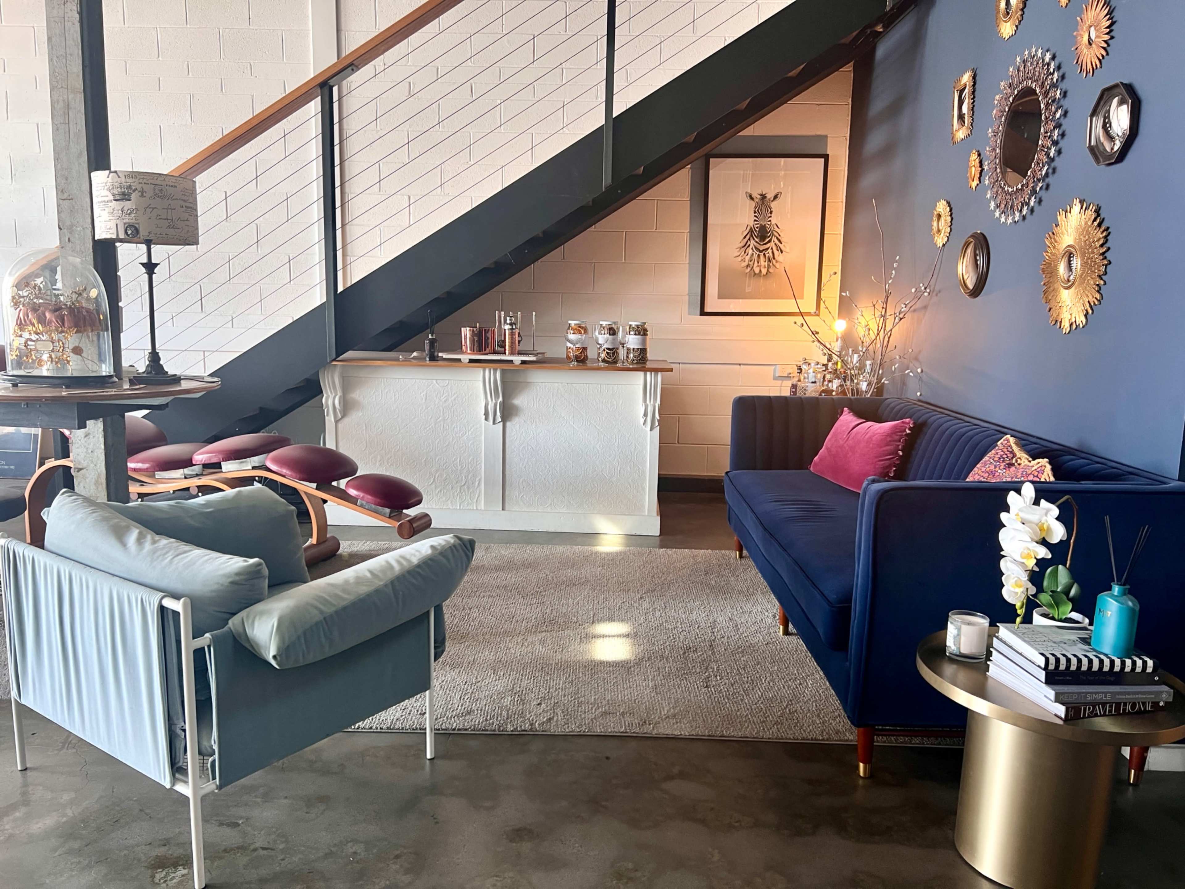 The image shows a modern lounge area featuring a navy blue sofa, a green chair, a decorative mirror wall, and a console table underneath a staircase.