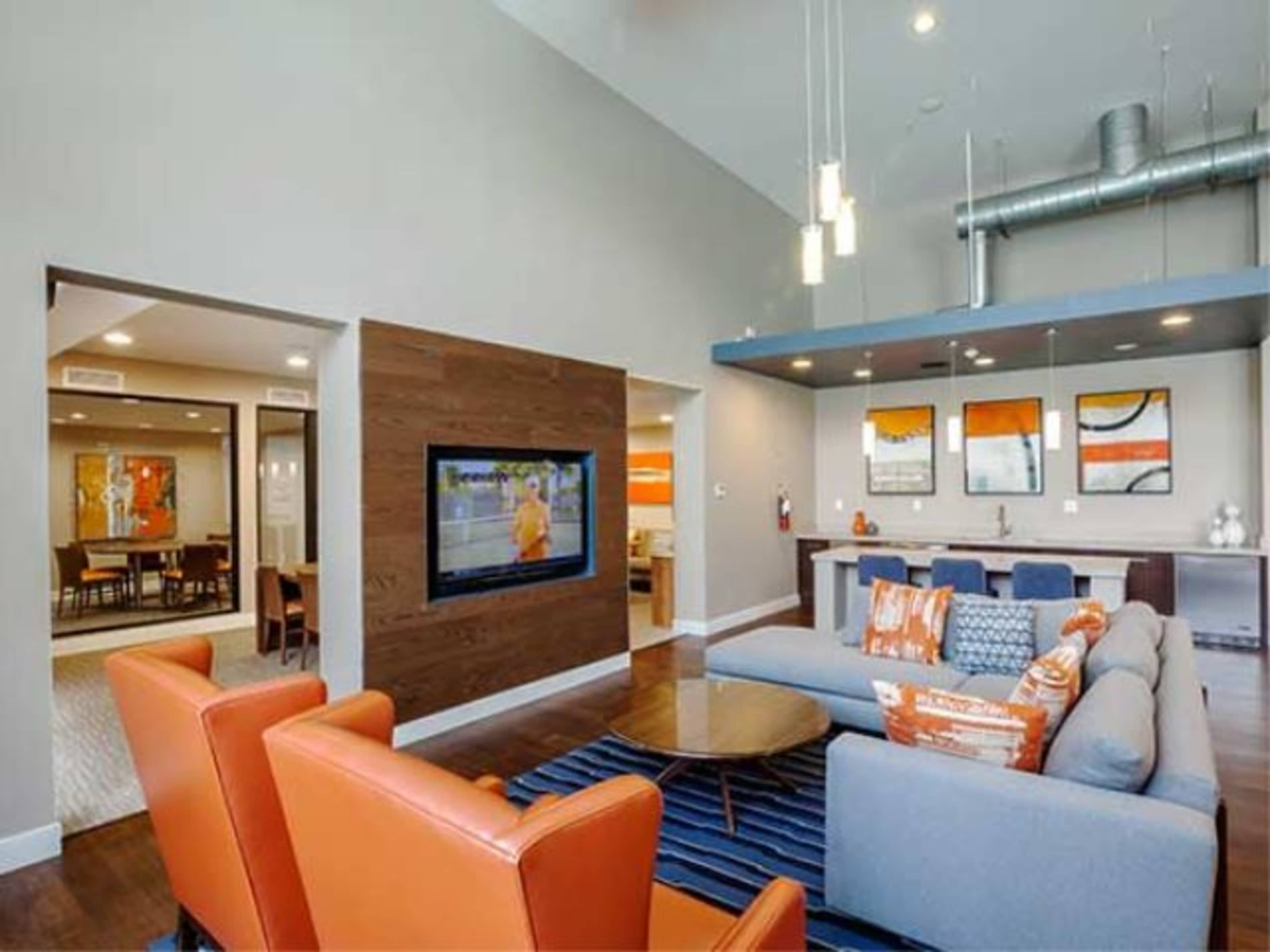 A modern common area with a living room featuring orange and blue furnishings, a TV on the wall, and a dining area in the background.