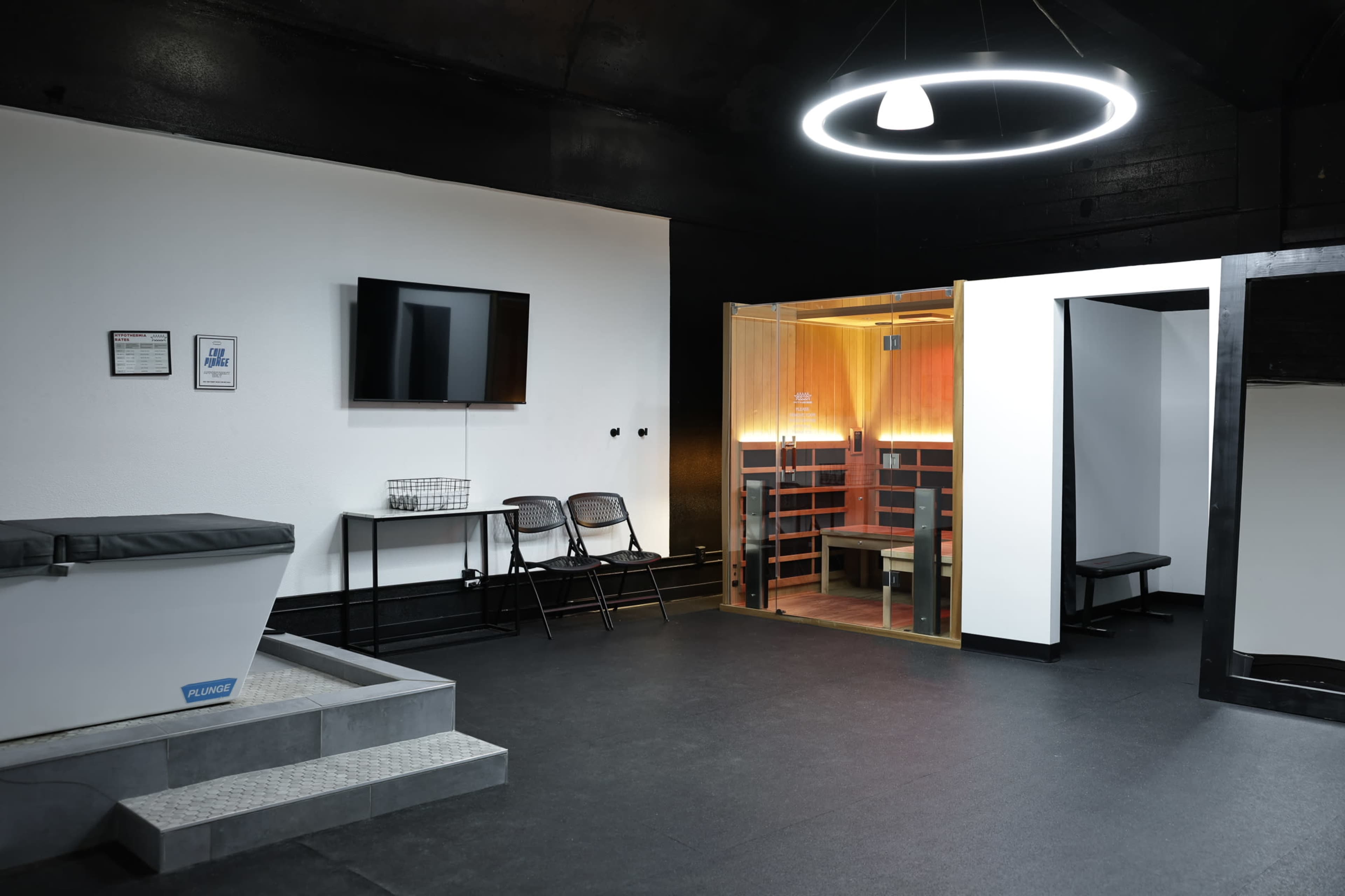 The image displays a modern wellness space featuring a wooden sauna, a small seating area with two chairs, a TV mounted on the wall, and a cold plunge tub.