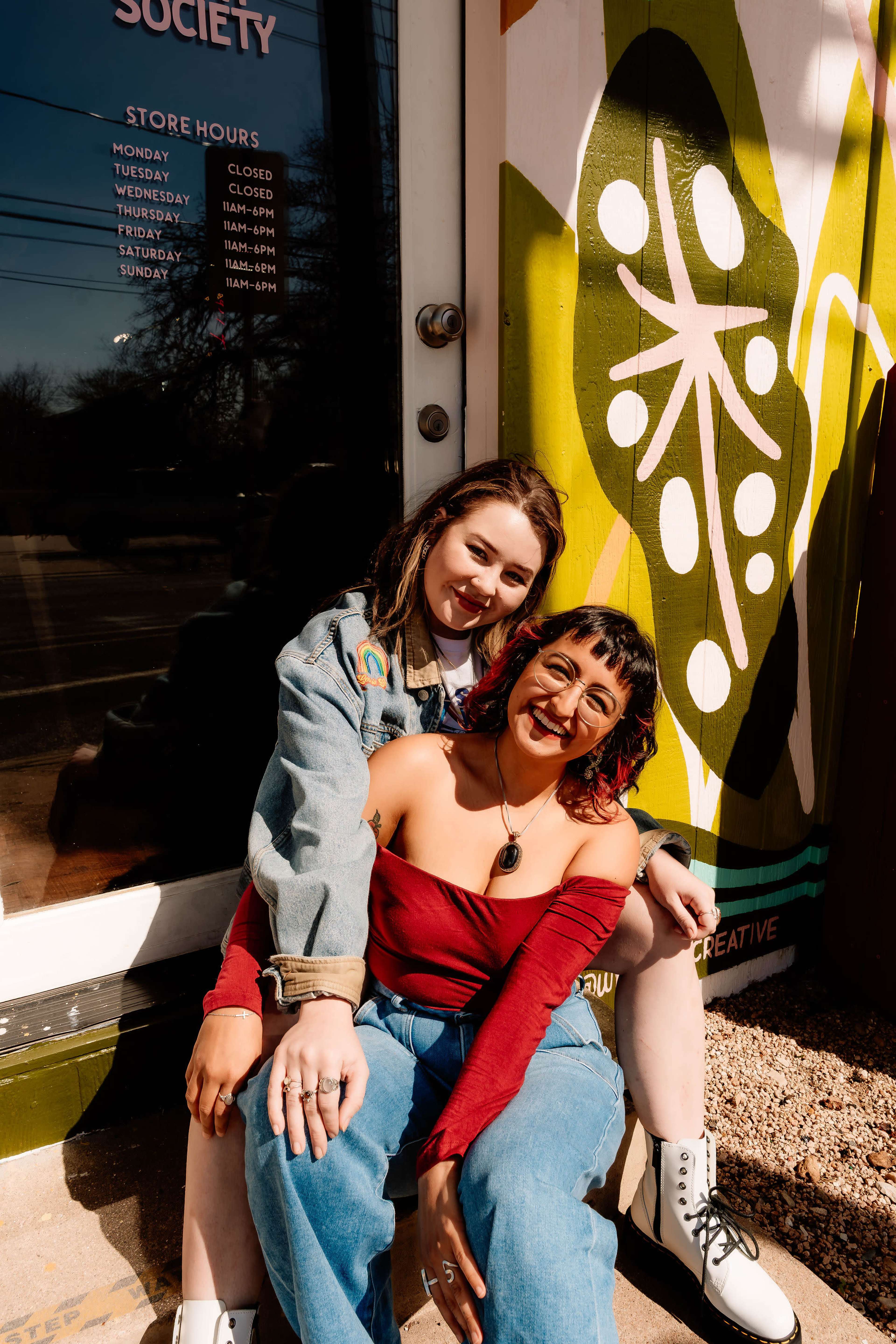 Two women sit closely together in front of a colorful mural and a storefront, smiling at the camera.