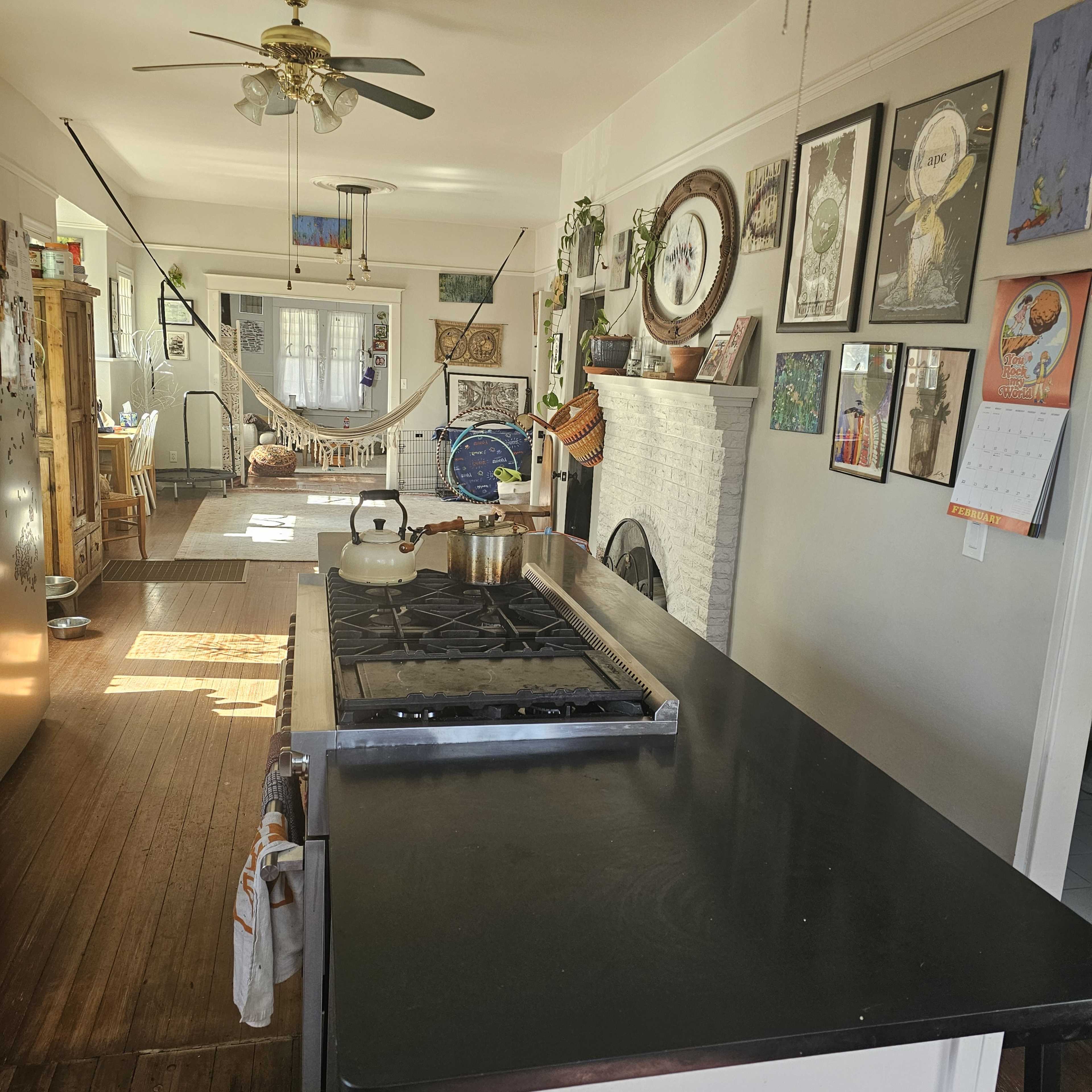 The image shows a kitchen with a black countertop, a stove with a kettle on it, and a view into a light-filled living area decorated with various artworks and plants.