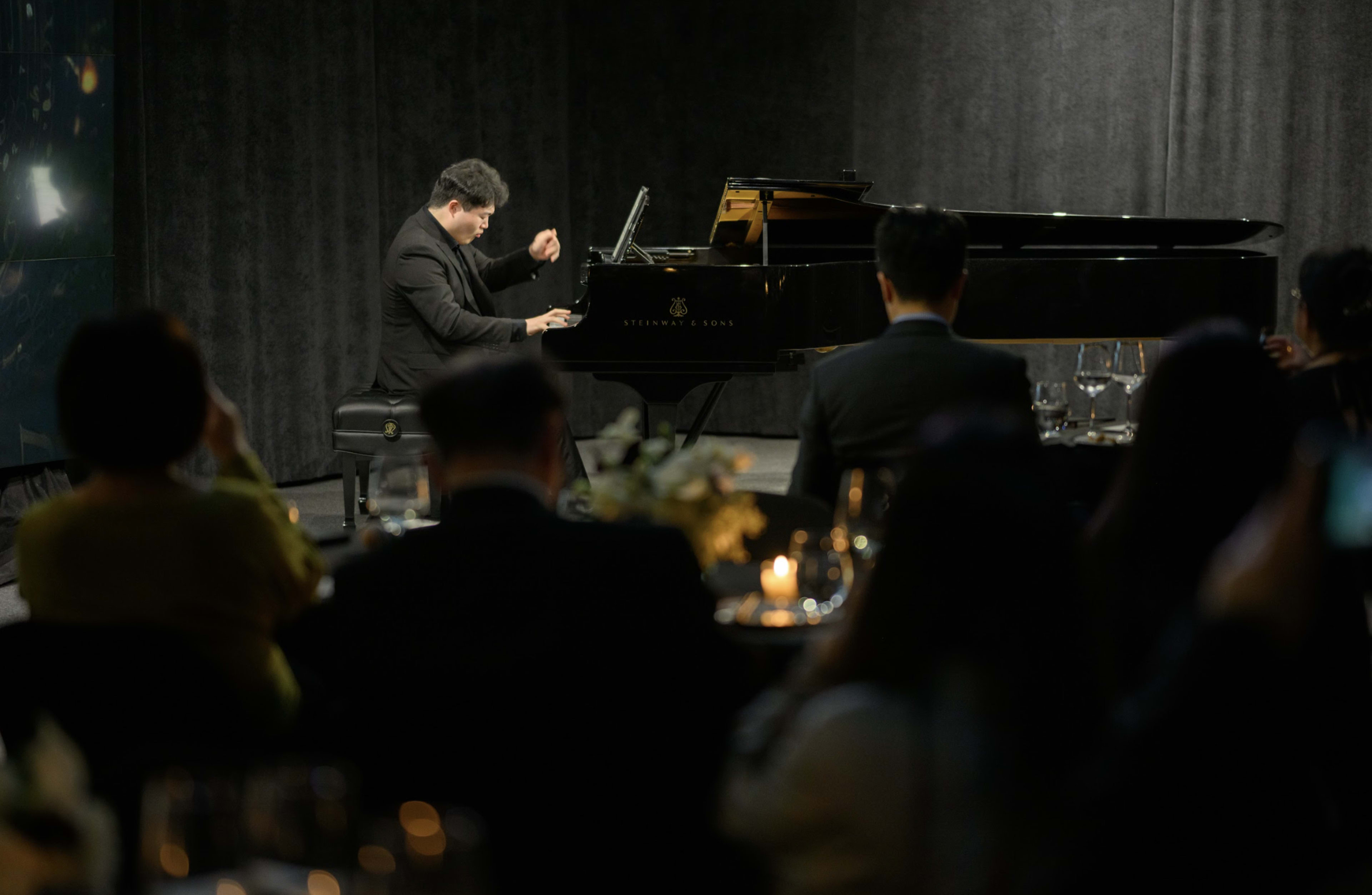 A pianist performs at a grand piano in front of an audience seated at elegantly set tables.