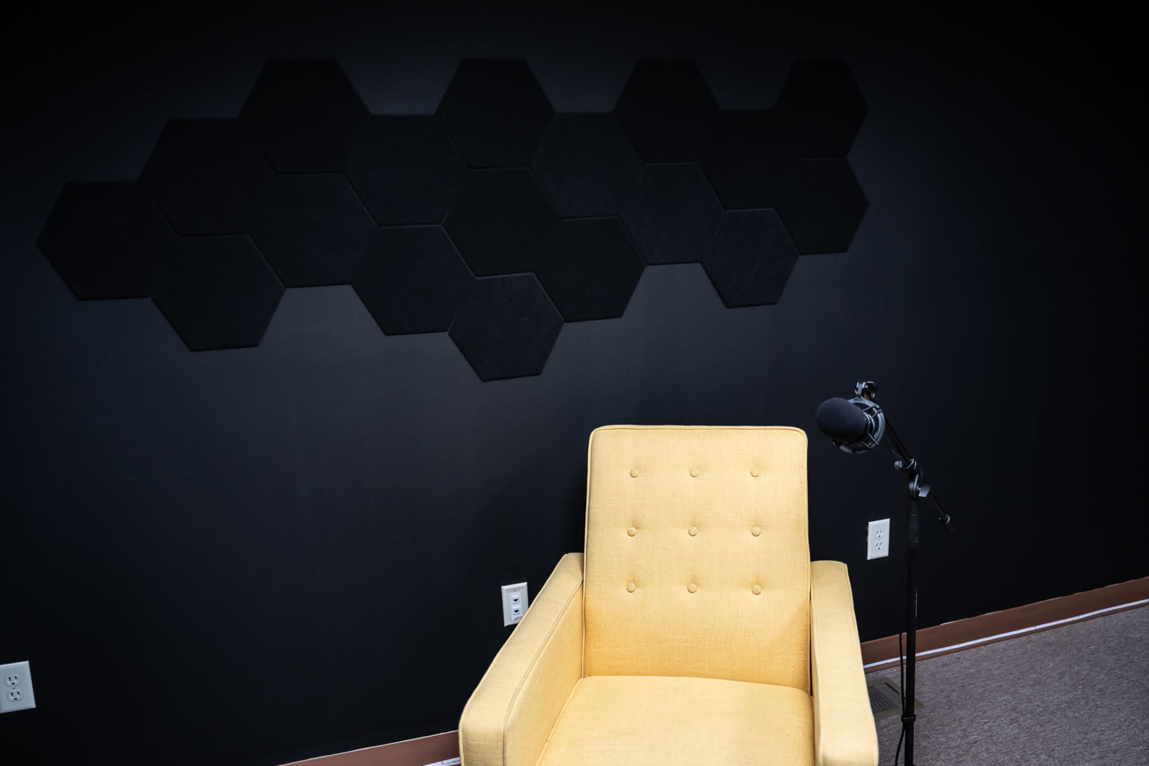 A yellow armchair is positioned next to a microphone on a stand in front of a black wall with hexagonal sound panels.