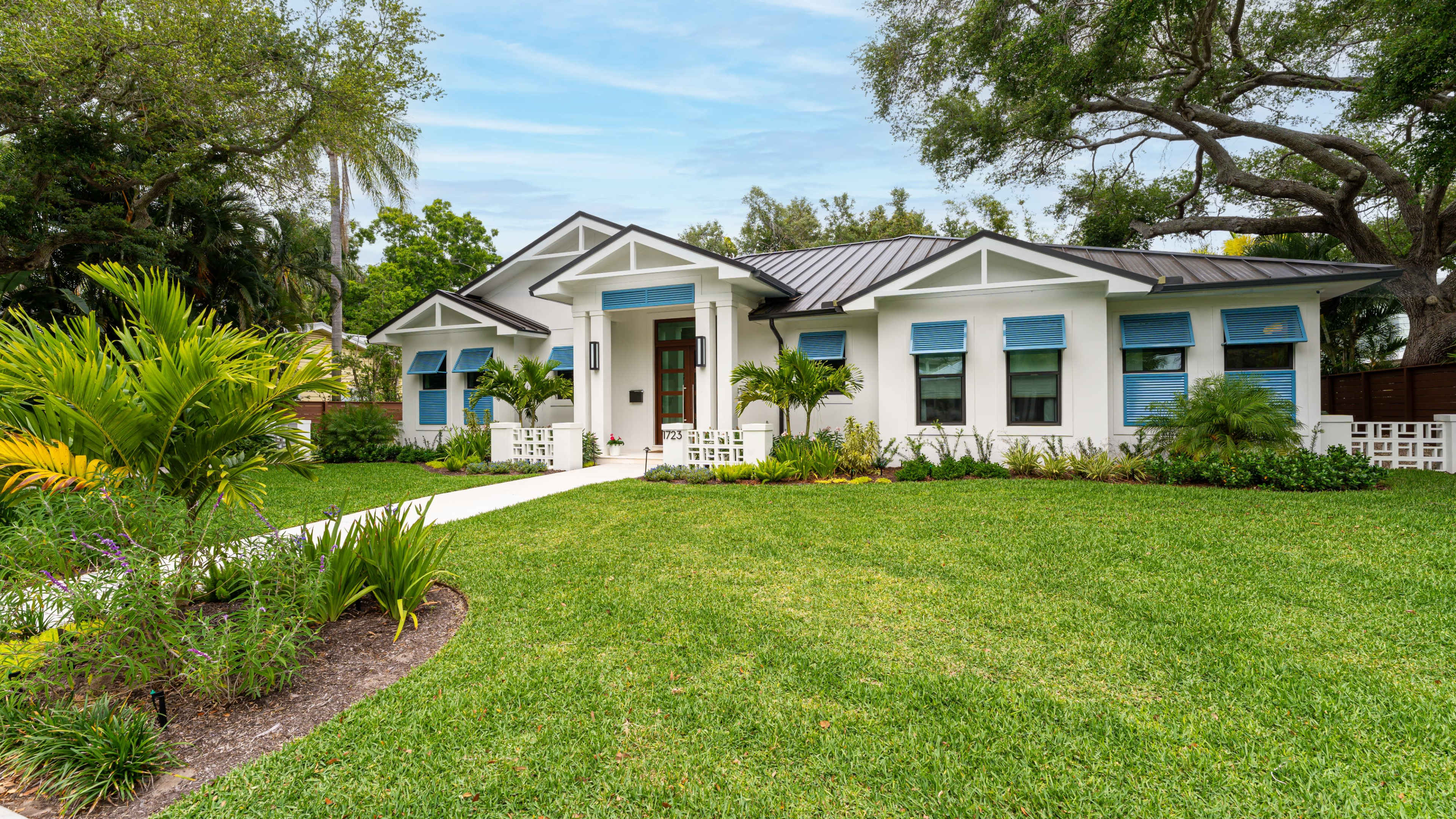 The image shows a single-story white house with a black roof, surrounded by lush greenery and well-maintained landscaping, featuring blue shutters on the windows.
