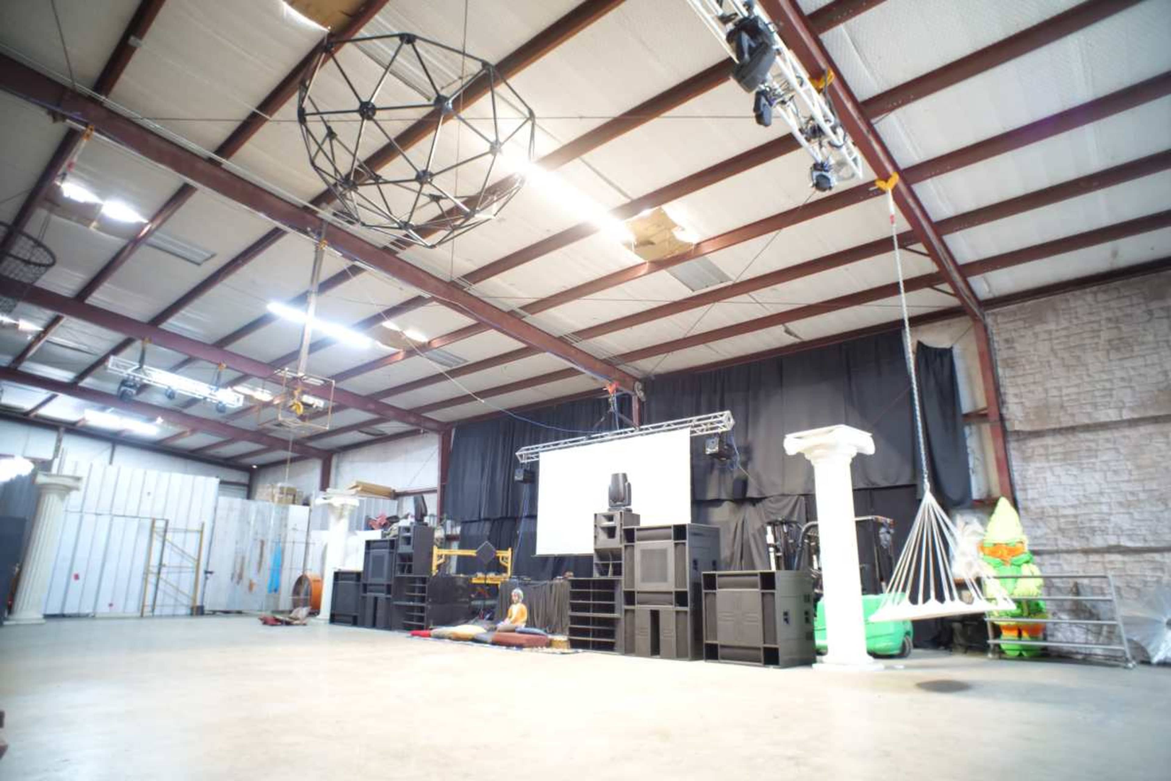 The image shows a spacious industrial interior with a raised stage, audio equipment, and various props arranged throughout the area.