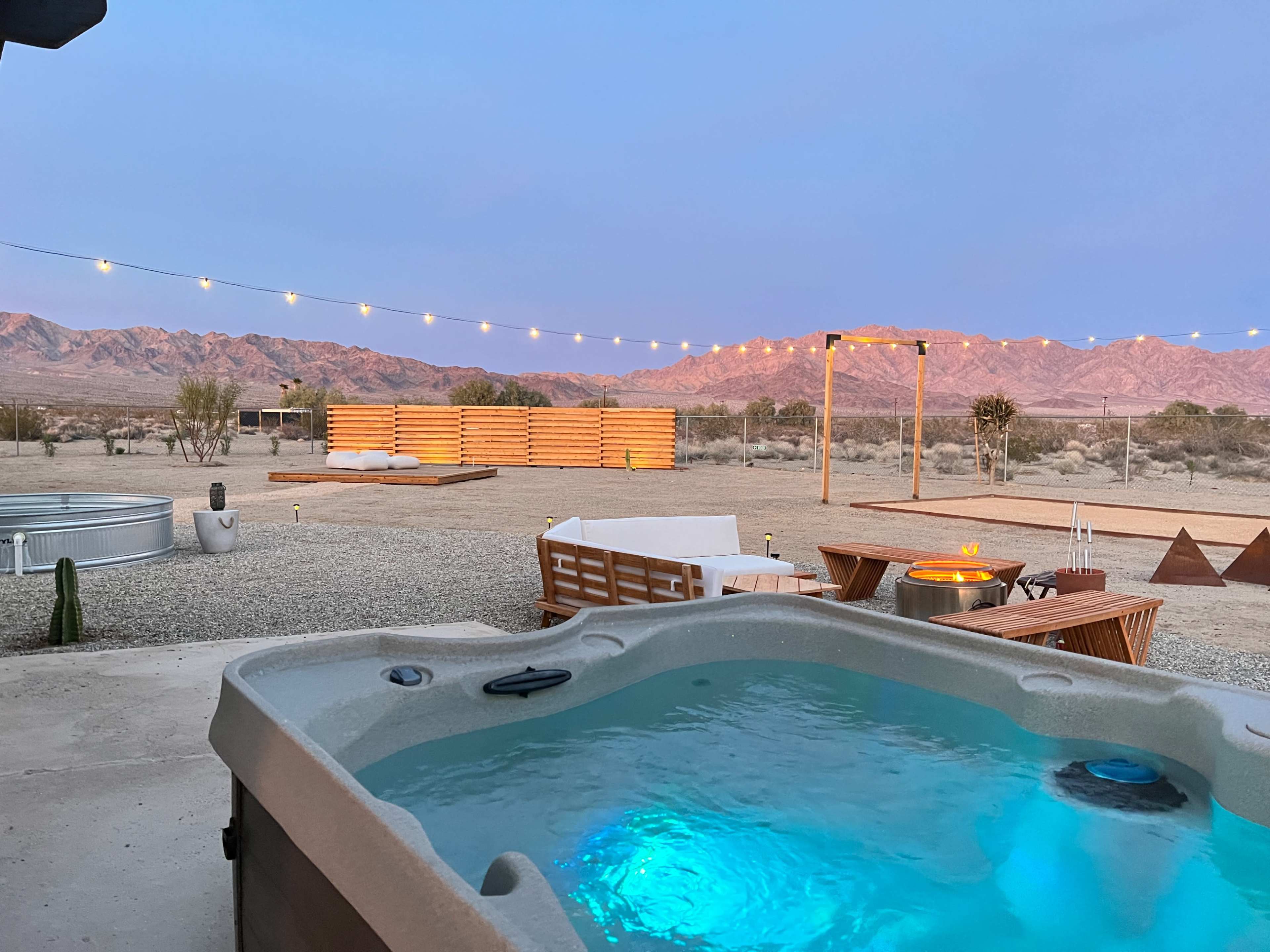The image shows a hot tub in the foreground with a desert landscape and mountains illuminated by sunset in the background.