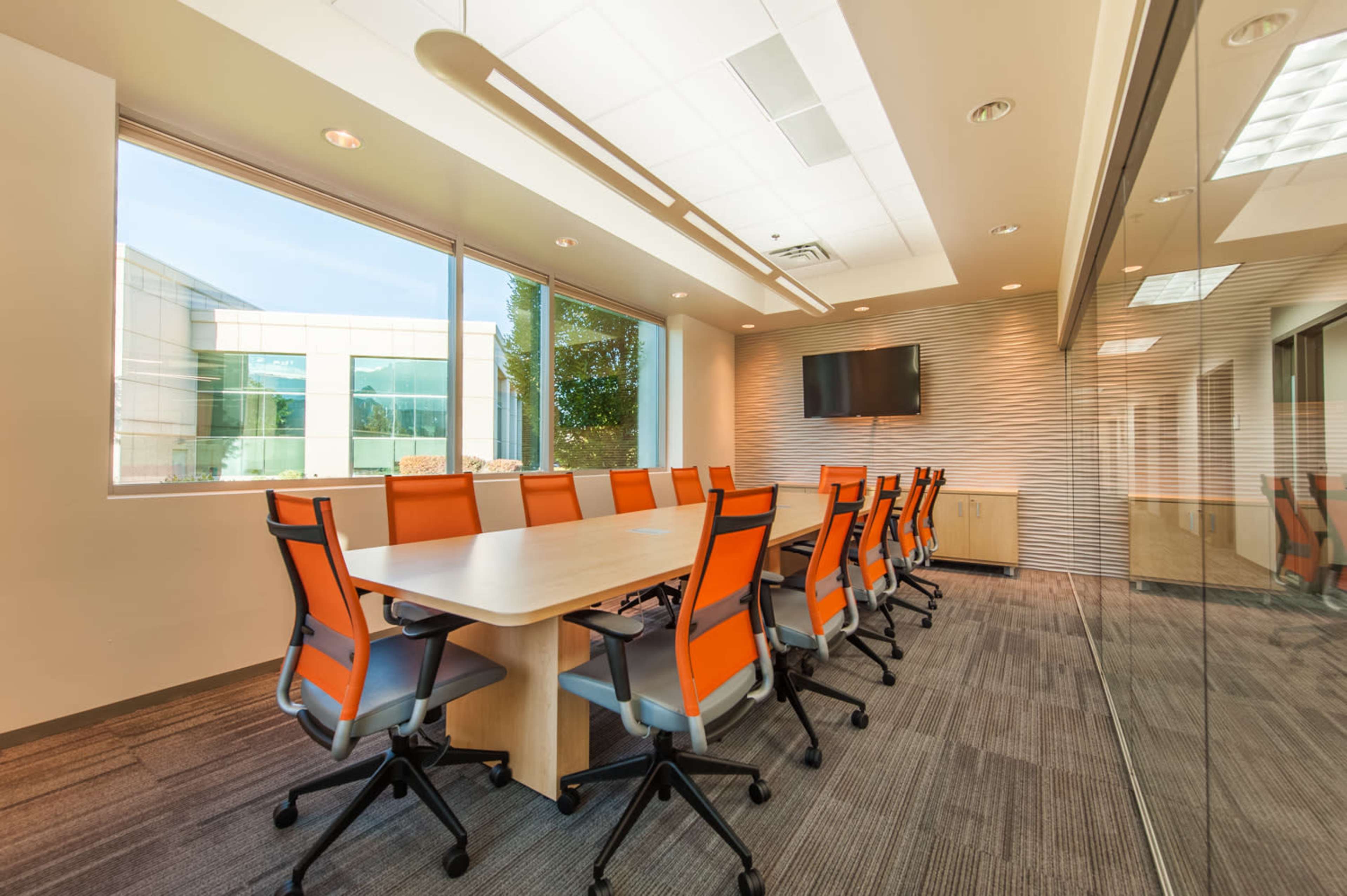 A conference room featuring a long table and orange chairs, with large windows that provide natural light and a view of the outside.
