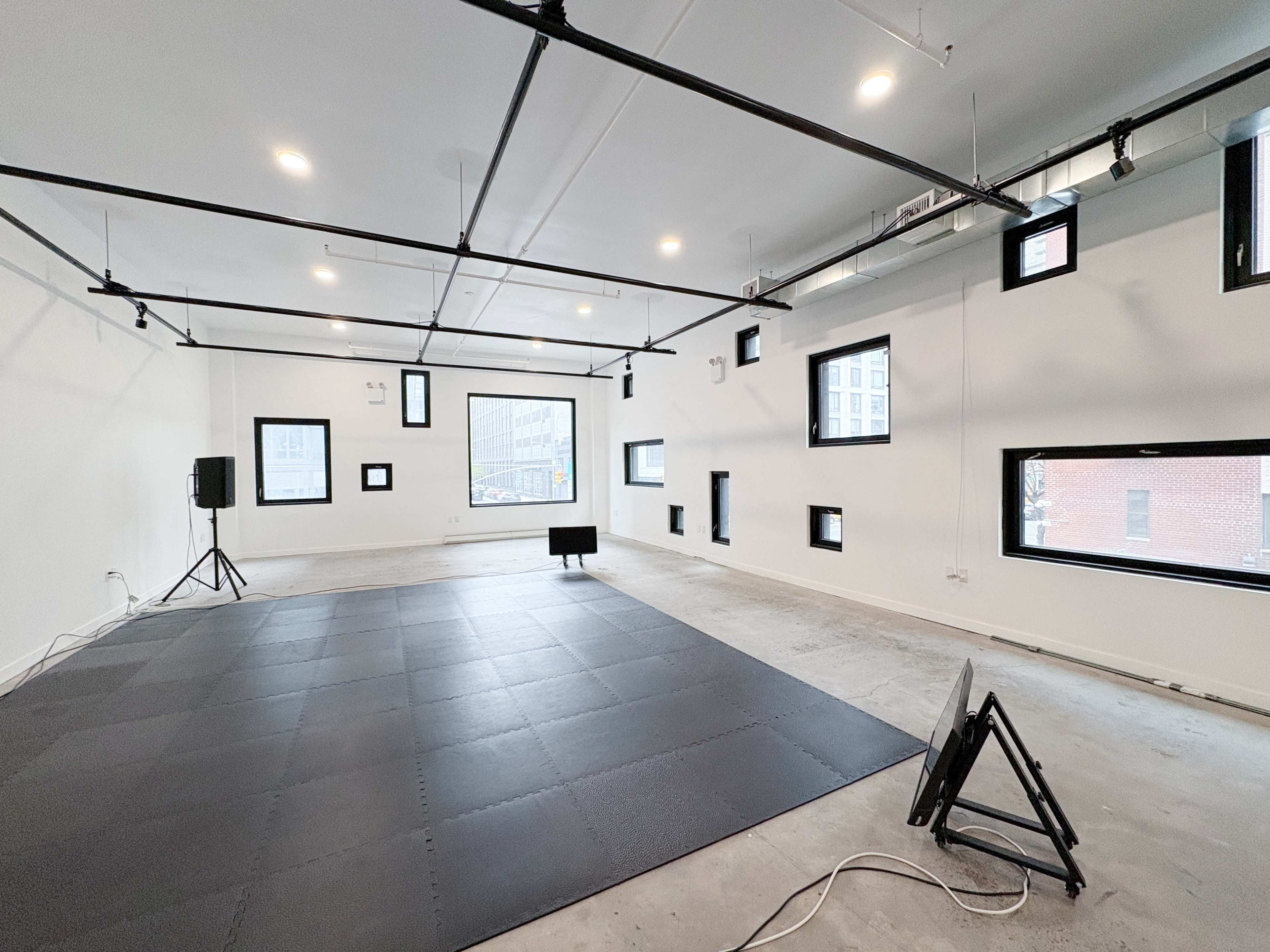The image shows a spacious, empty room with a black mat on the floor, large windows, and visible industrial-style fixtures on the ceiling.