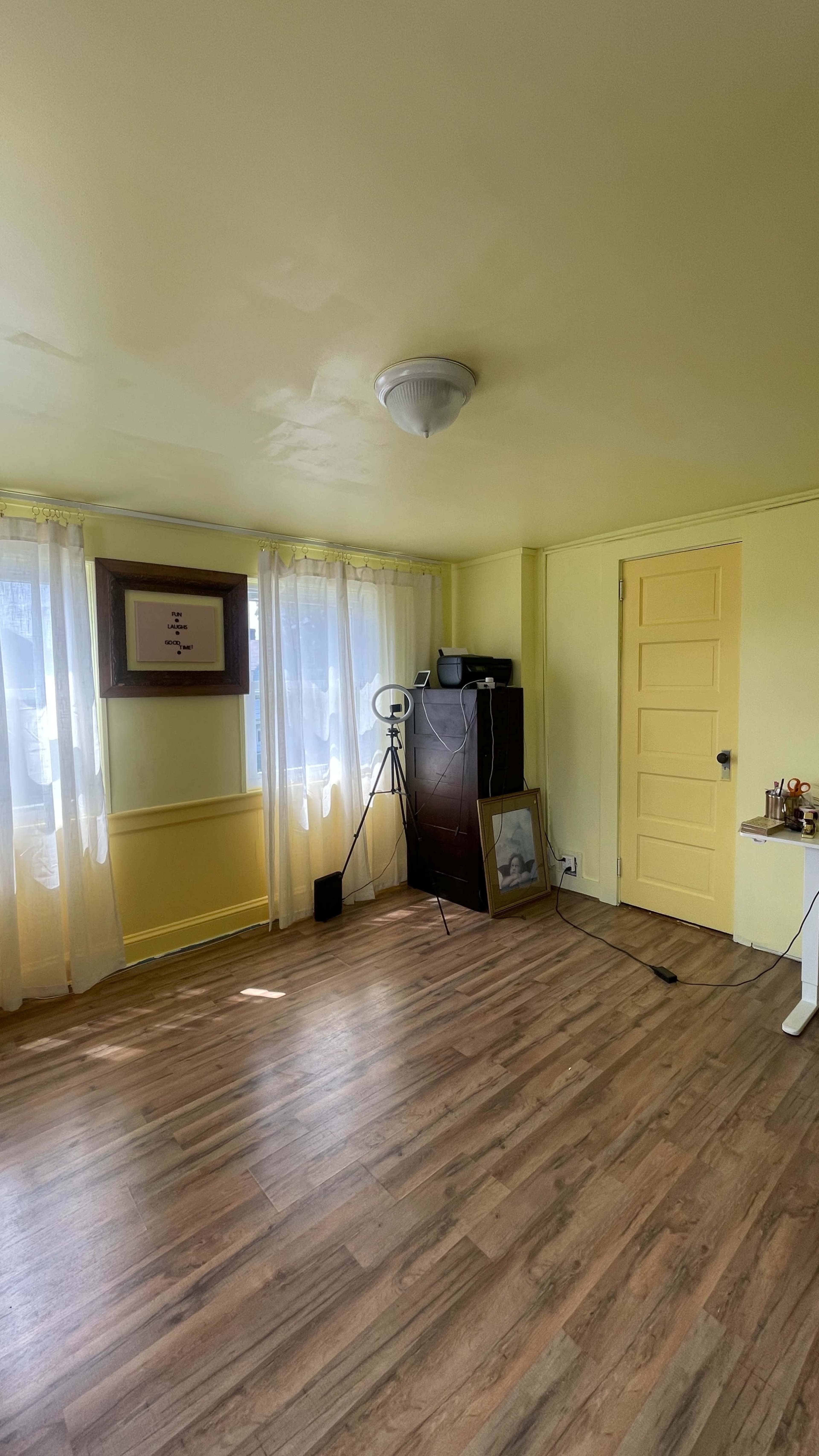The image shows a yellow-walled room with wooden flooring, featuring two large windows with sheer curtains, a door on the right, and a few pieces of furniture including a standing mirror and a small table.
