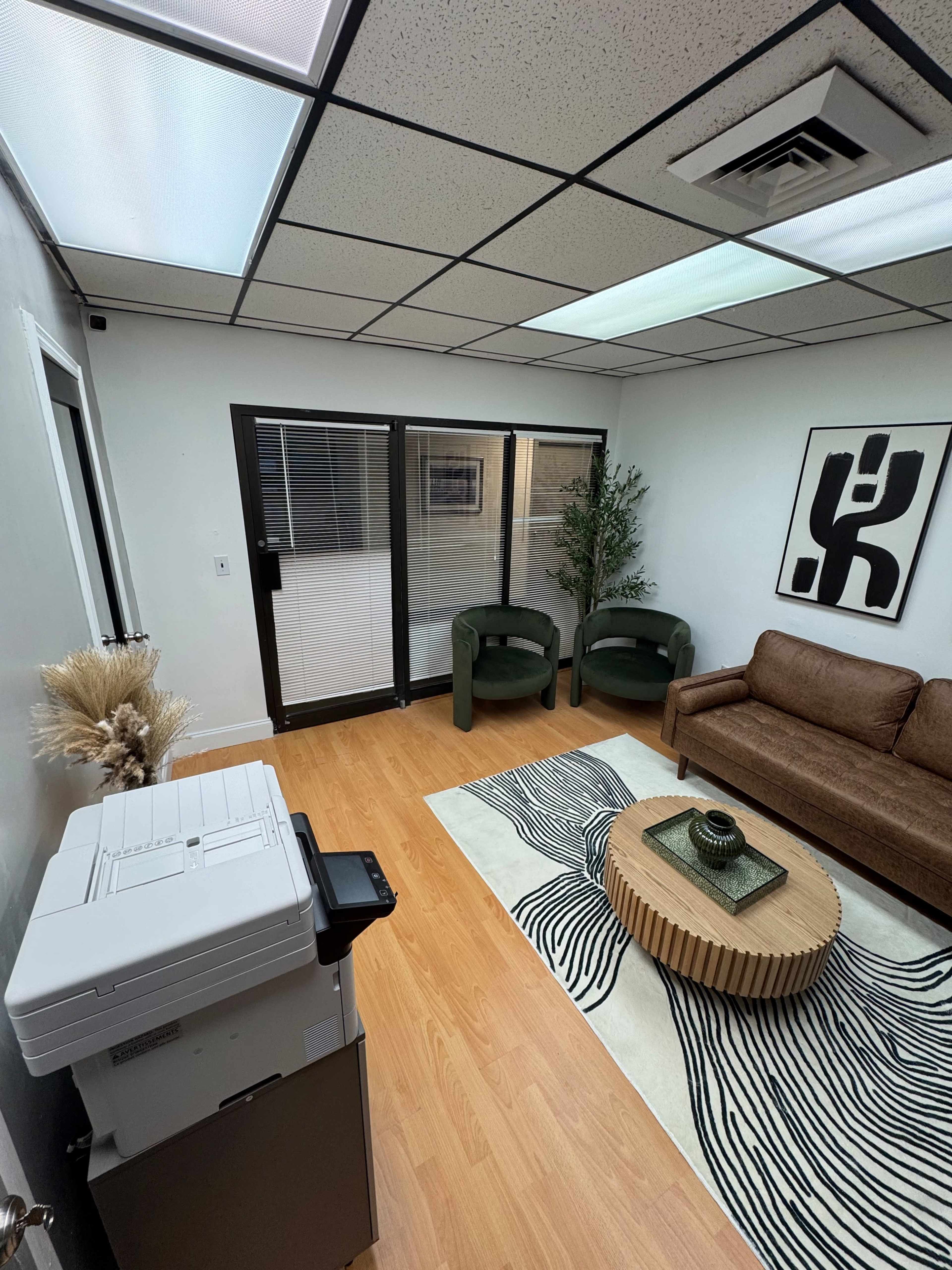 The image shows a small office room with a printer, two green chairs, a brown sofa, a round coffee table, and large windows letting in natural light.