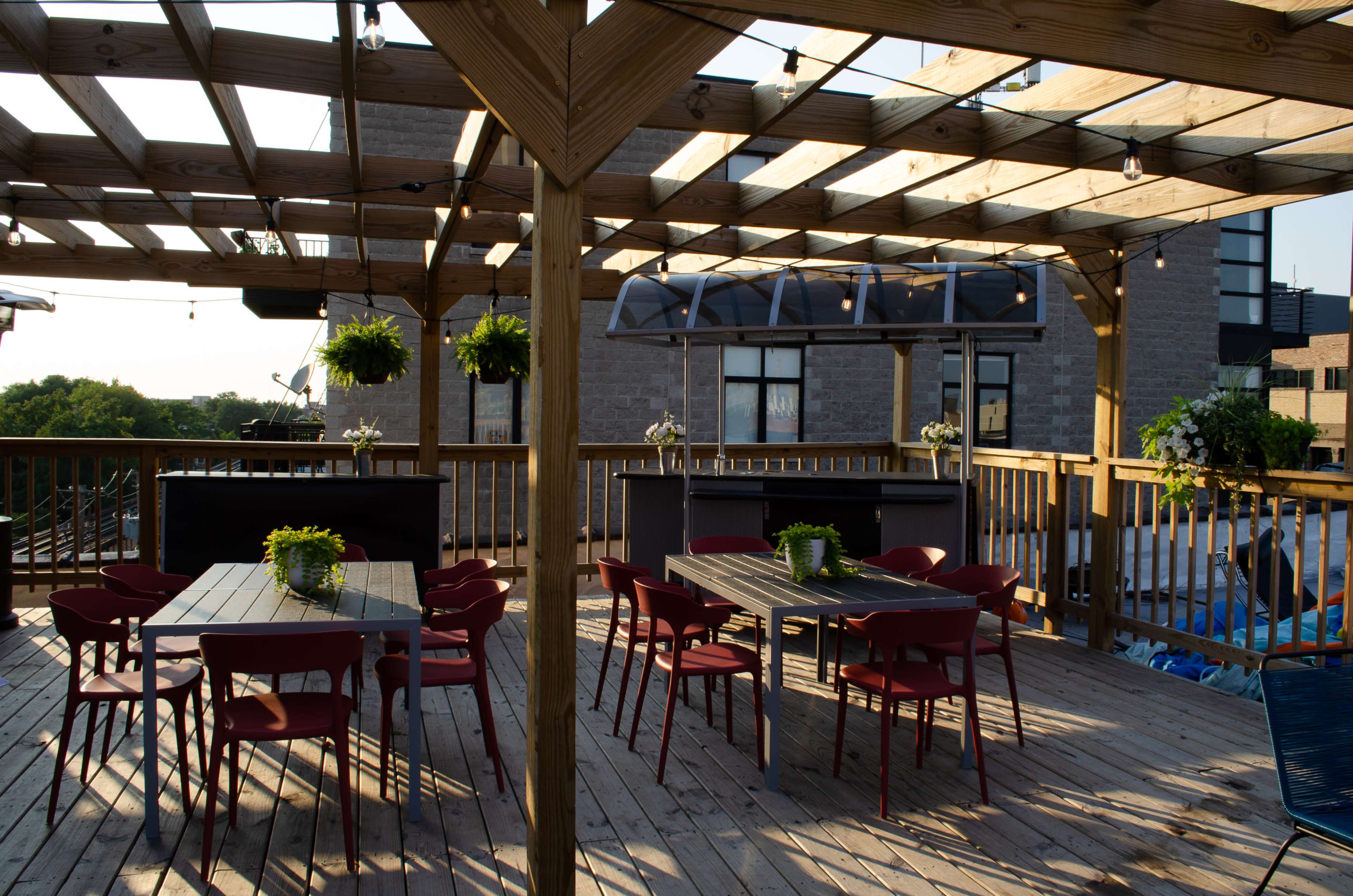 A wooden rooftop deck with several tables and chairs, surrounded by plants and string lights, under a clear sky.