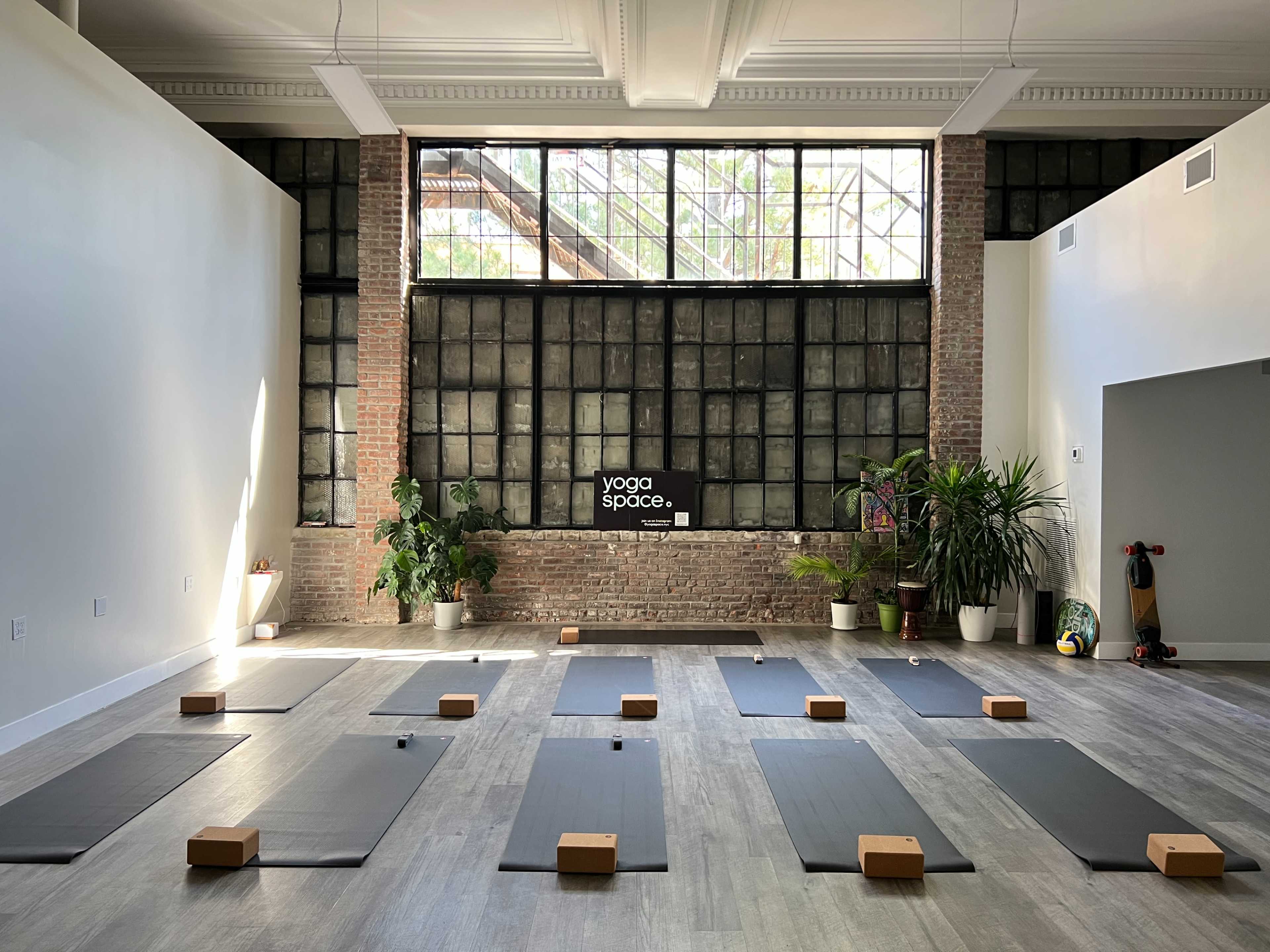 A spacious yoga studio with mats arranged on the floor, wooden blocks in front of each mat, and large windows letting in natural light.