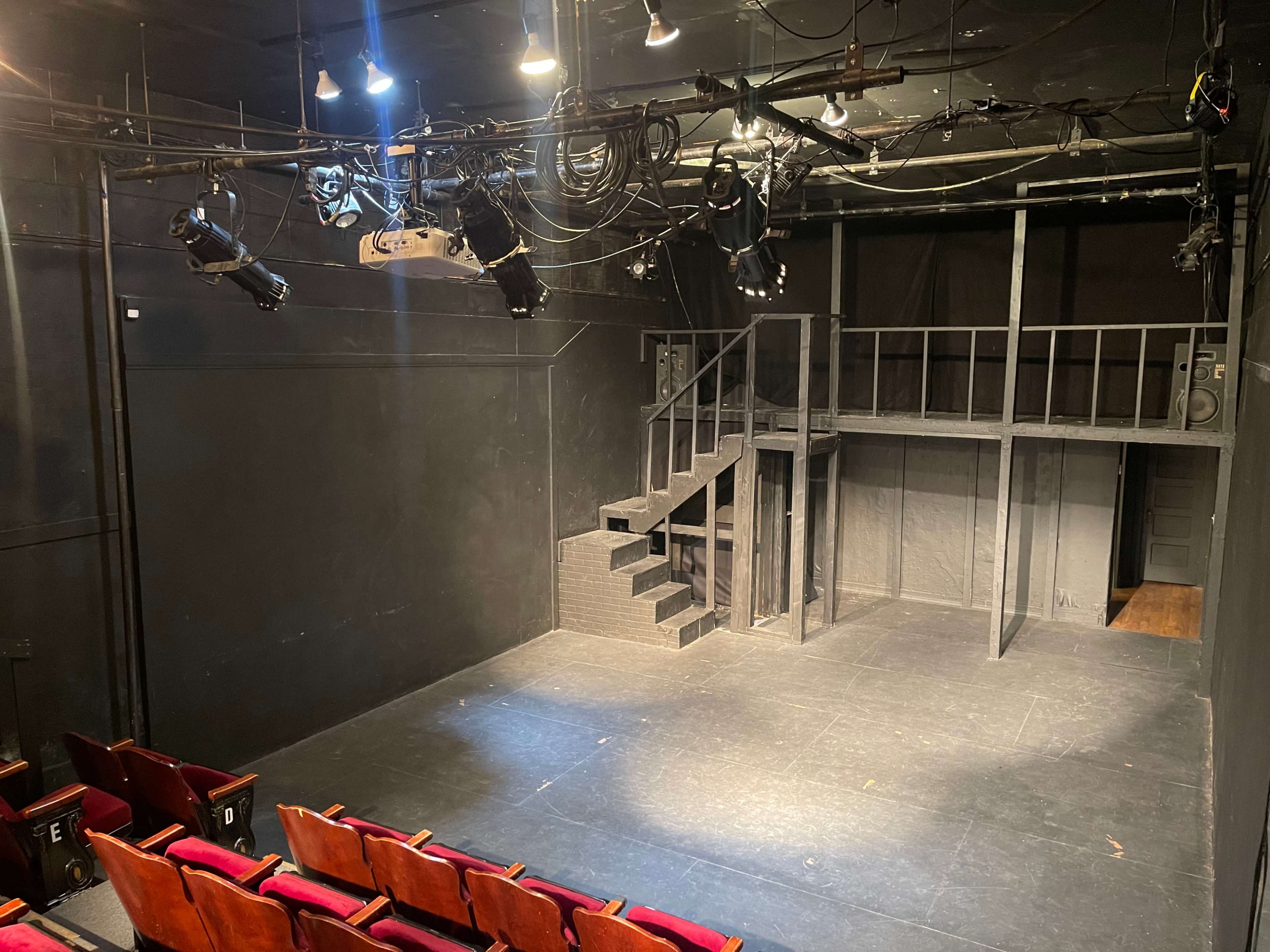 A dimly lit black box theater with a raised platform, stairs, and empty red seats facing the stage.