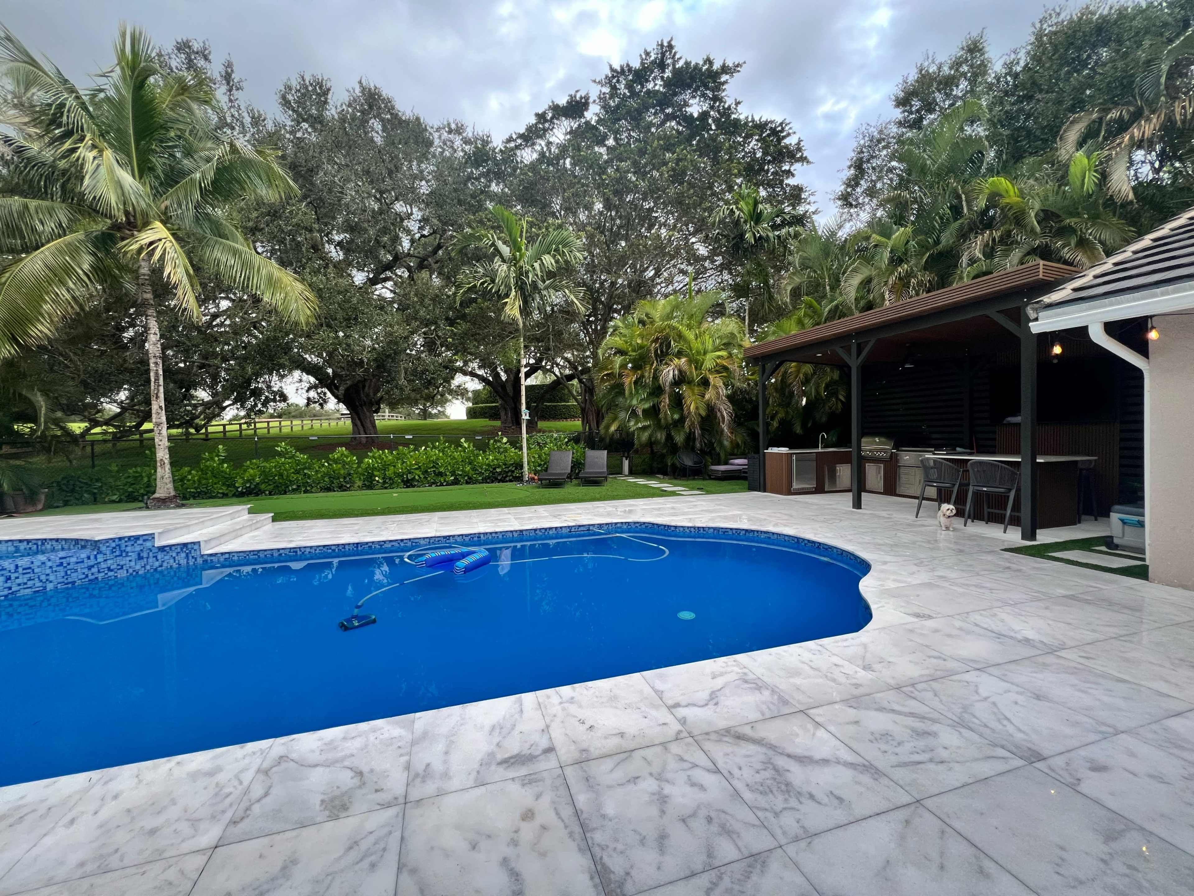 The image shows a residential backyard with a swimming pool surrounded by tropical plants and a covered outdoor seating area.