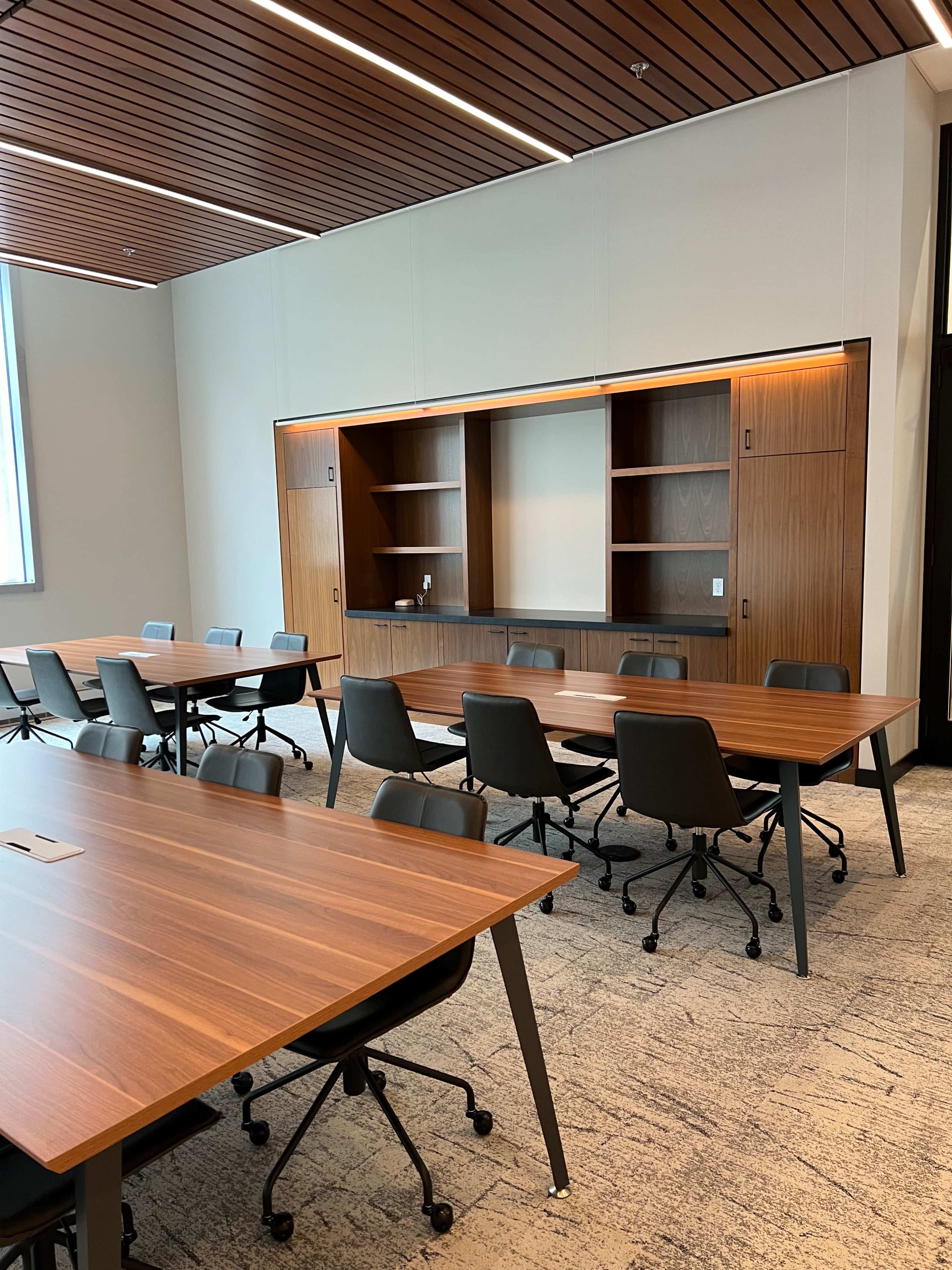 A modern conference room features several rectangular wooden tables and rolling chairs arranged around a centrally located built-in shelving unit.