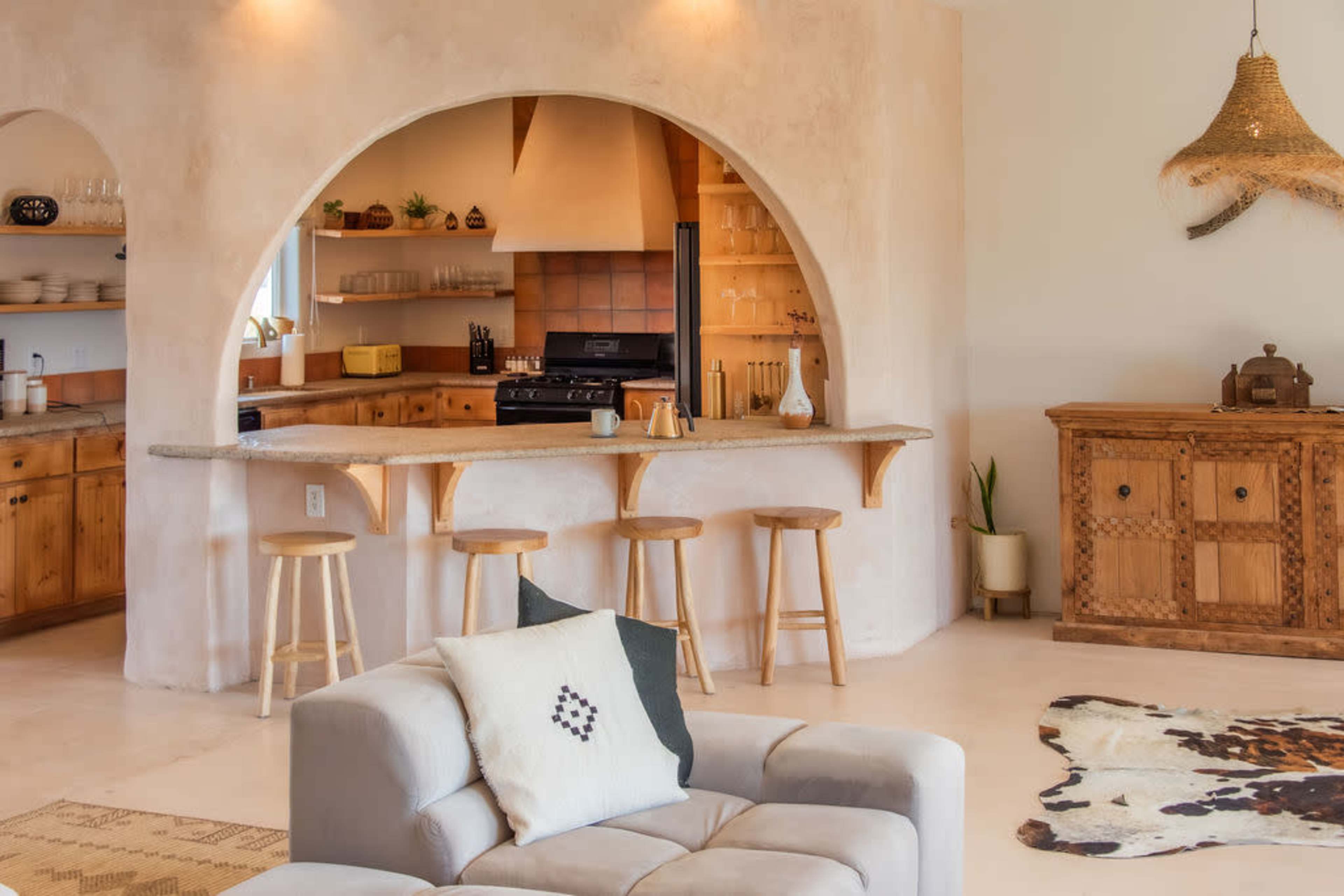 The image shows a modern kitchen with a curved archway, wooden cabinetry, and a breakfast bar with stools, all set in a bright, open space.