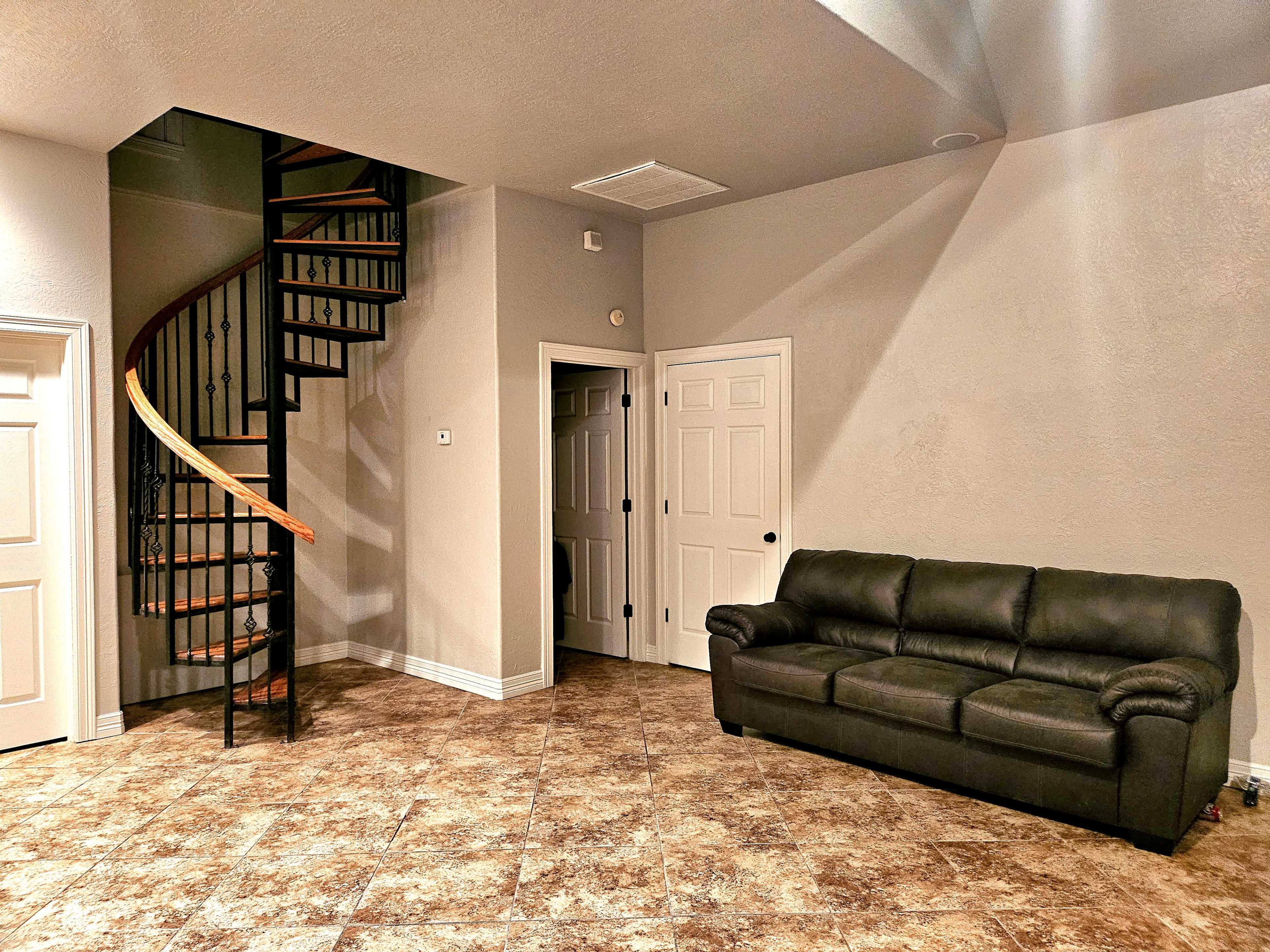 The image shows a living area featuring a black couch, a spiral staircase in the corner, and two white doors leading to separate rooms.
