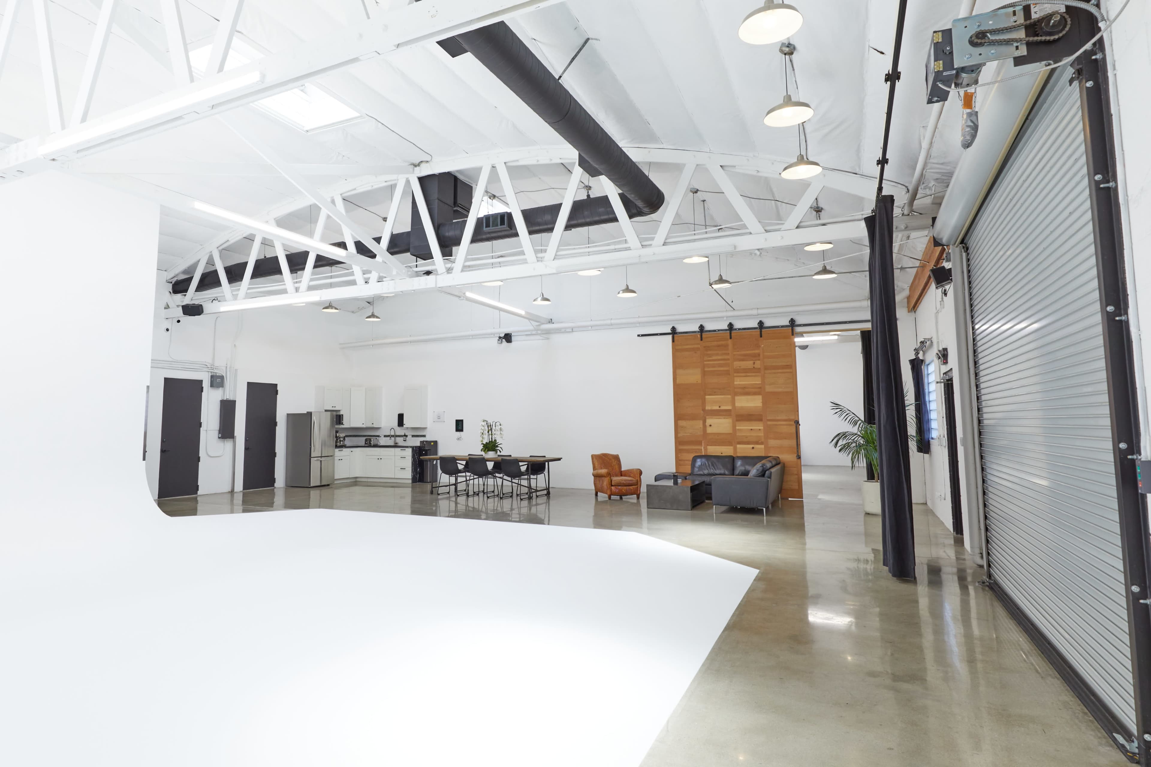The image shows a spacious and modern studio with a seamless white backdrop, a kitchen area, a dining table, and seating arrangements.