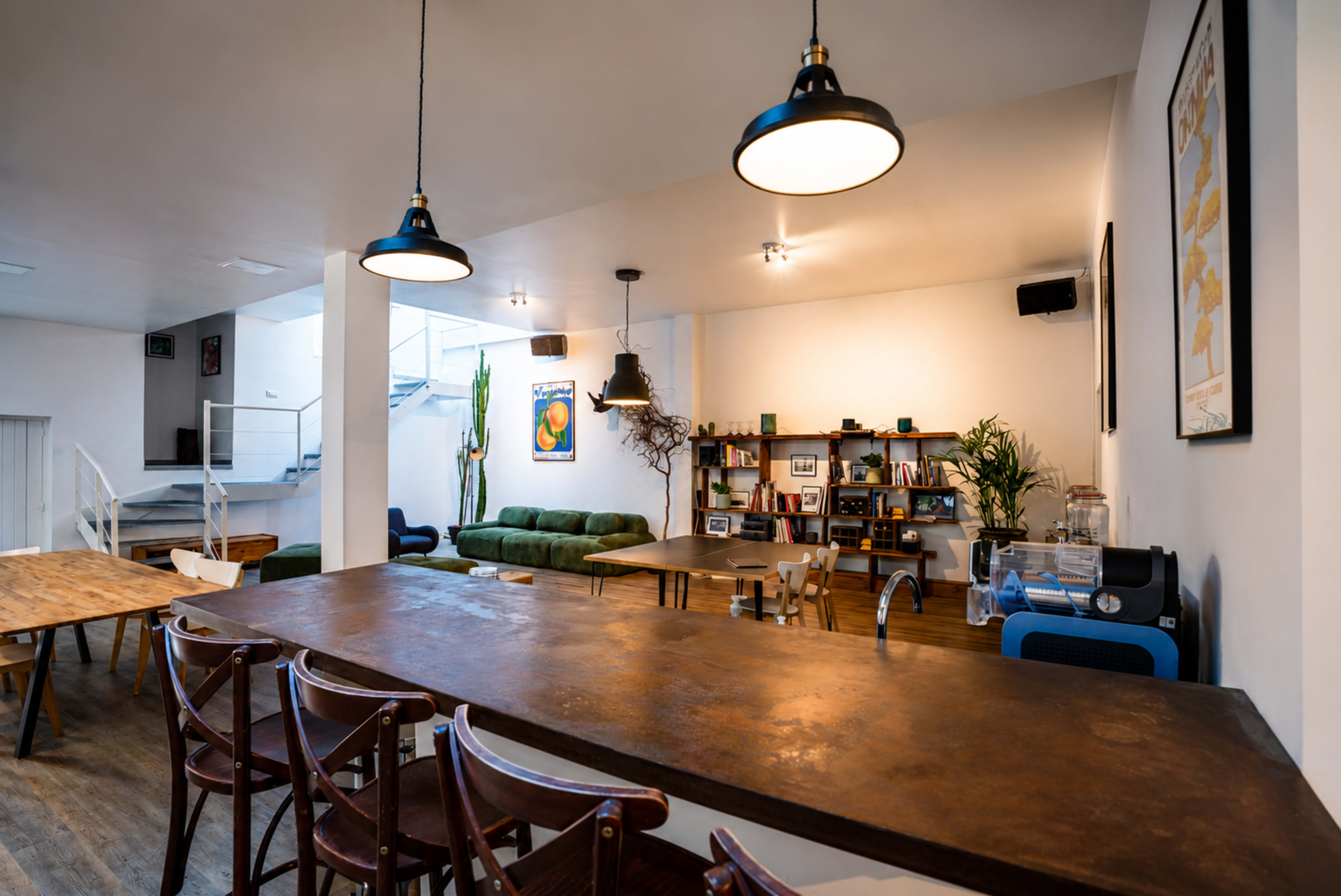 The image shows a spacious, modern interior of a café or co-working space with a large wooden table, green sofa, and shelves filled with books and plants.