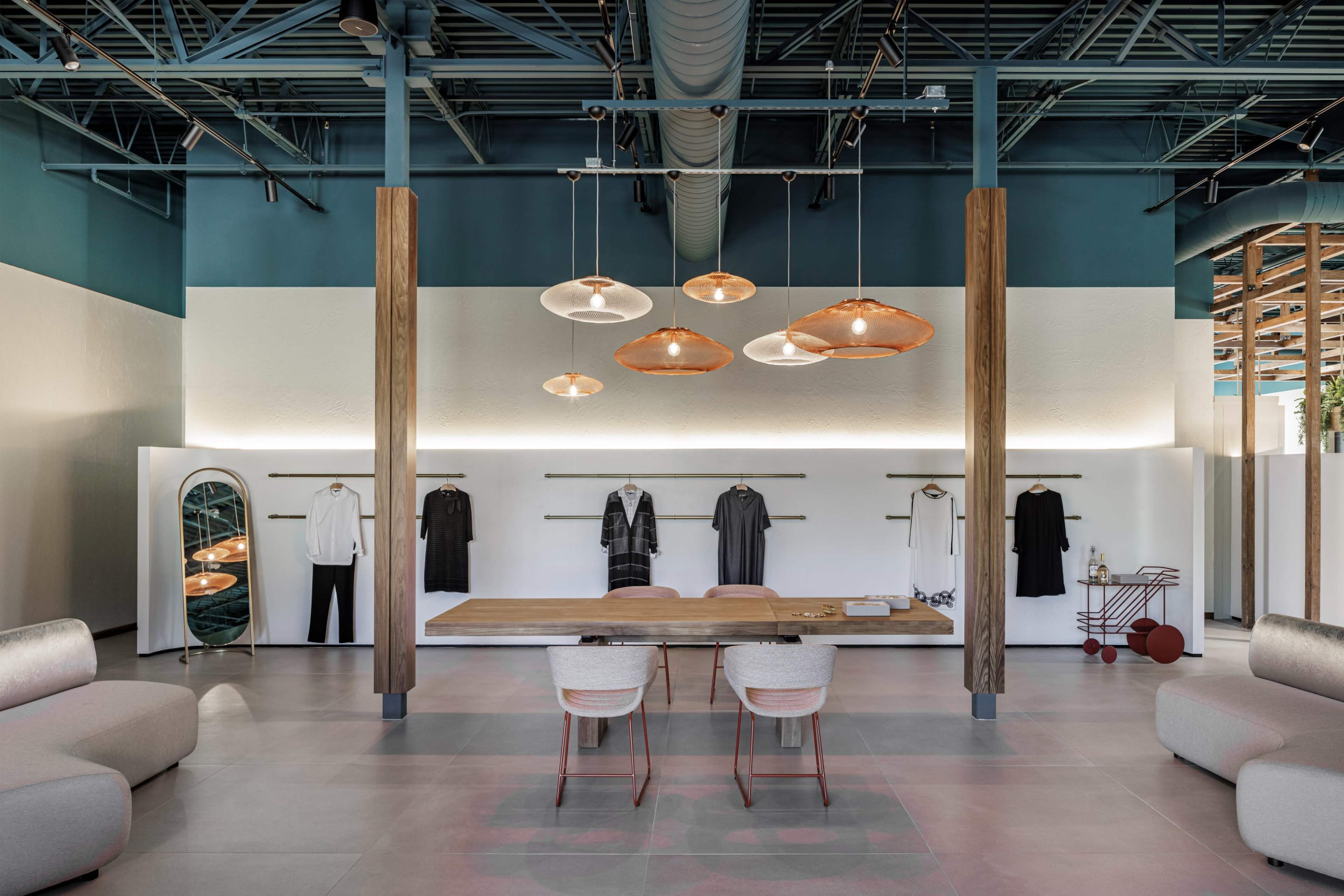 The image shows a modern retail space featuring a central table with chairs, clothing racks displaying garments, and contemporary pendant lights above.