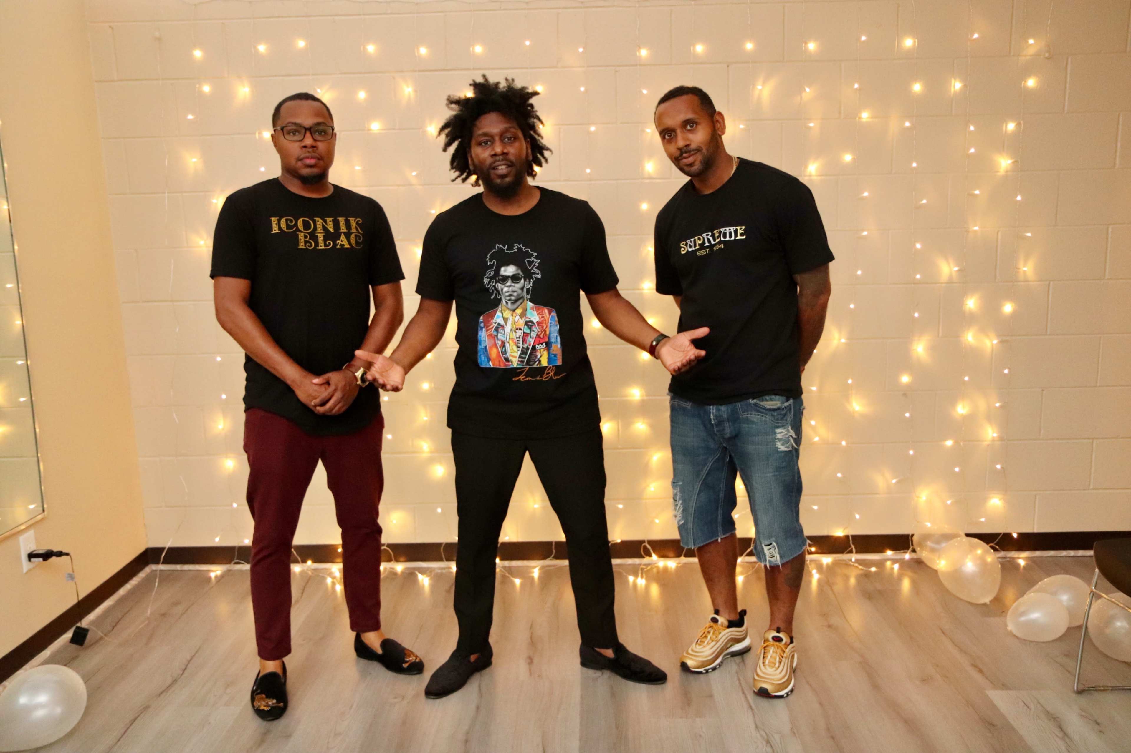 Three men stand in front of a wall adorned with string lights, wearing casual black t-shirts, with two in fitted pants and one in shorts.