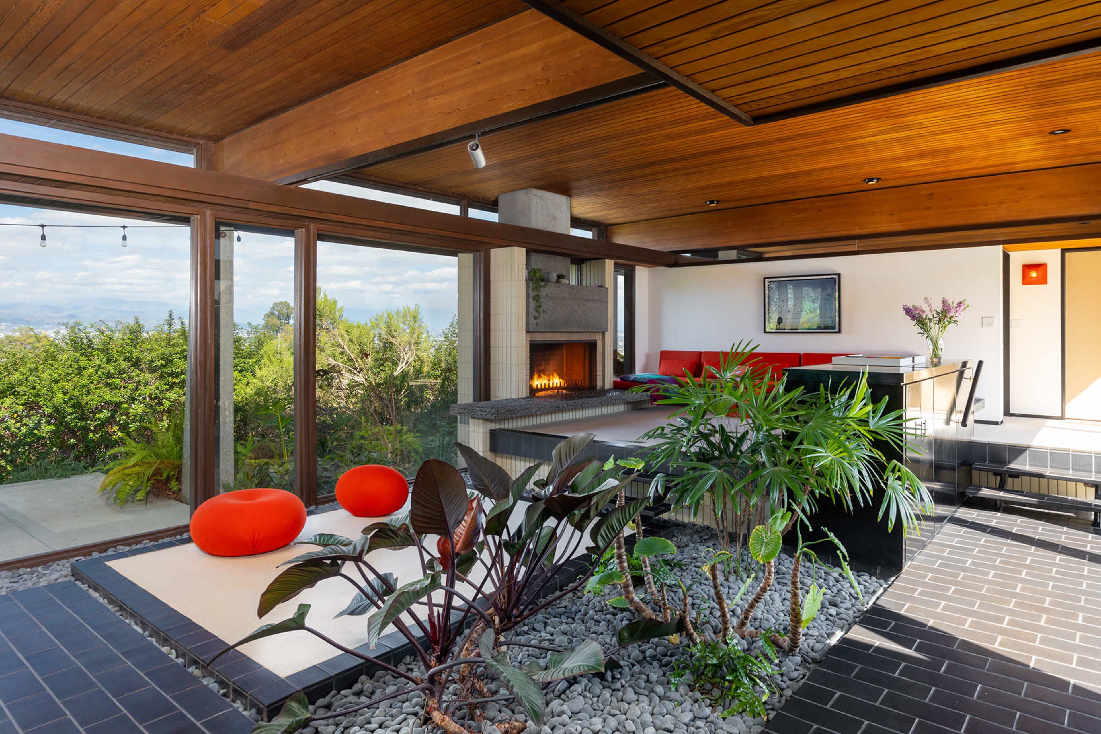 The interior of a modern home features wooden ceiling beams, a fireplace, and planters with lush greenery, alongside contemporary seating areas.