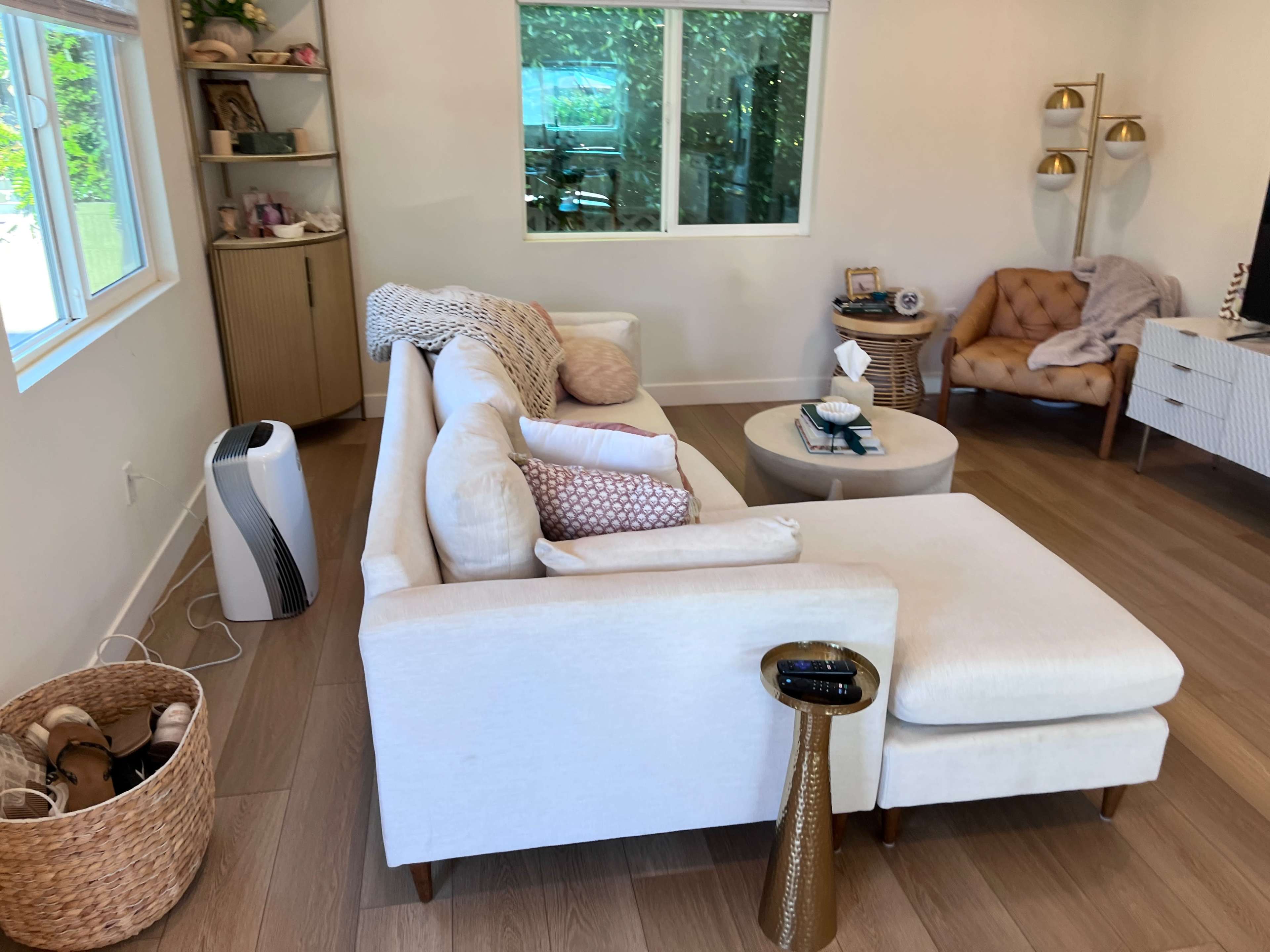 The image shows a modern living room with a white sectional sofa, a round coffee table, and a basket on the floor, along with various decorative elements and a large window.