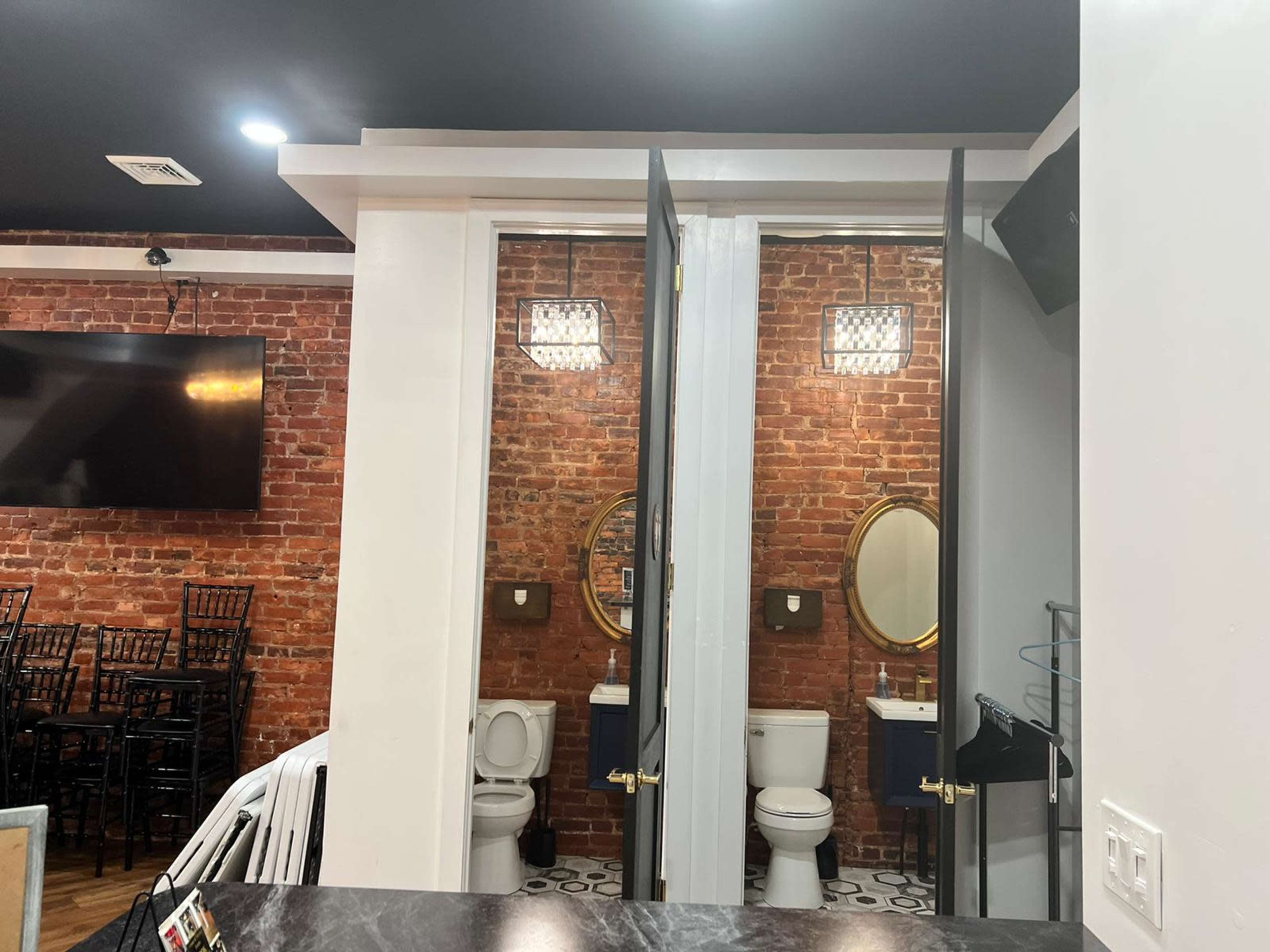 A restroom area featuring two stalls with large mirrors, surrounded by exposed brick walls and modern lighting fixtures.