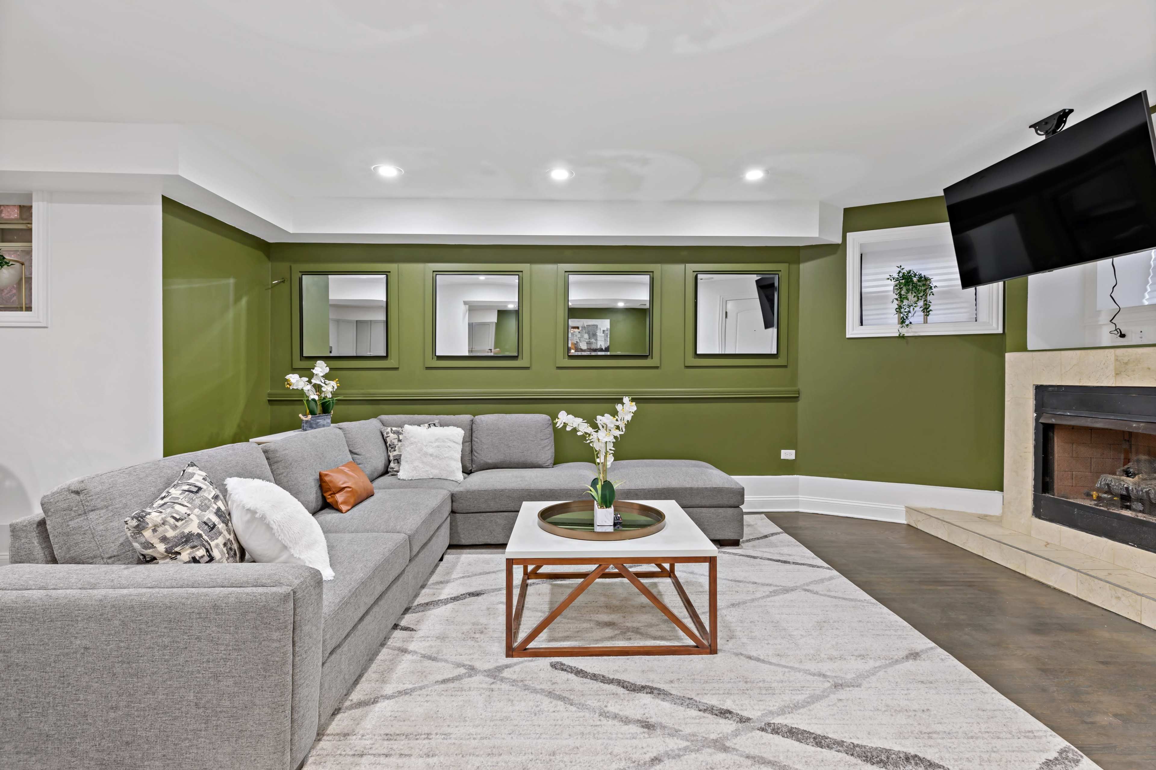 The living room features a sectional gray sofa, a white coffee table with a wooden base, a fireplace, and green walls with mirrors and plants.