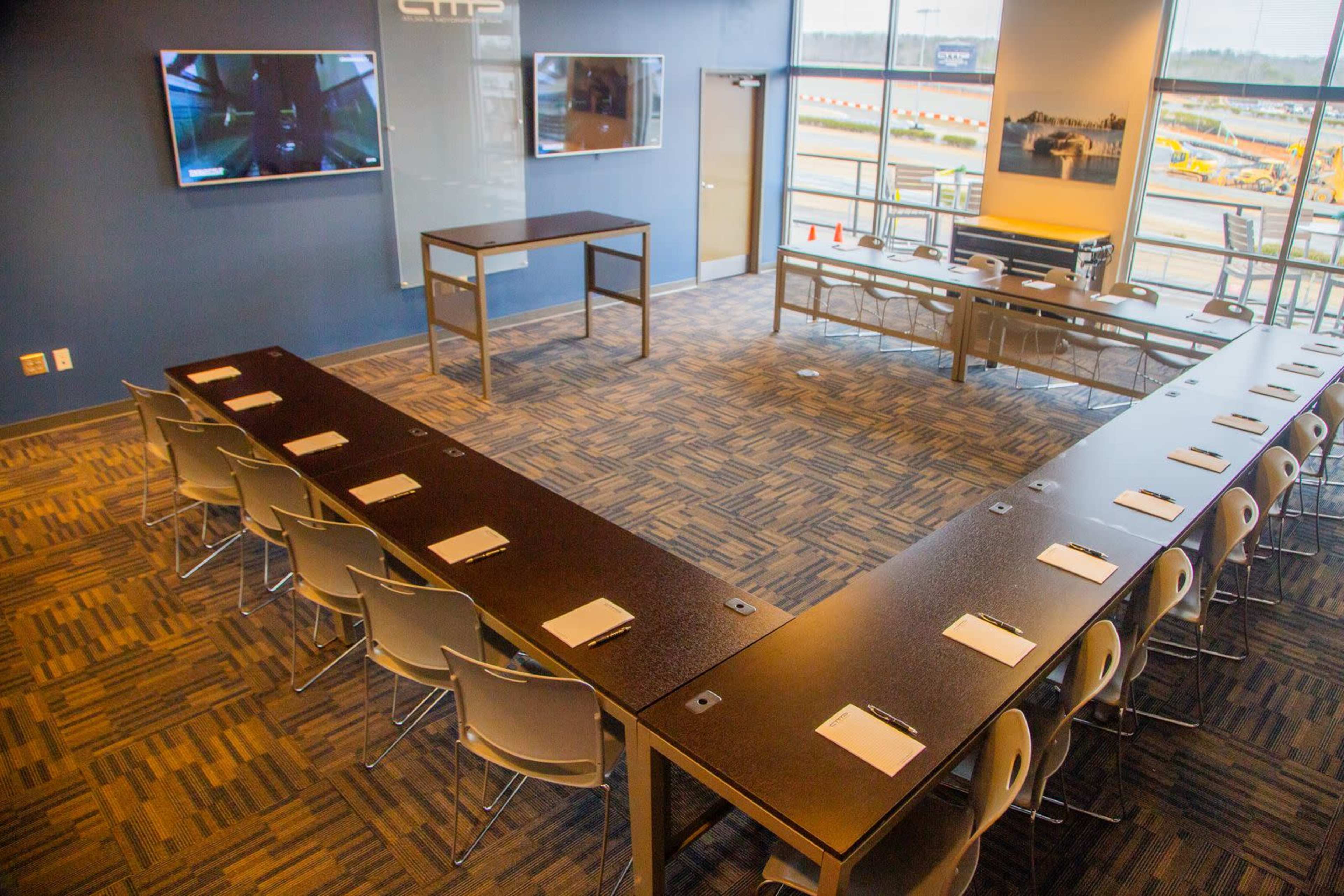 The image shows a conference room arranged in a U-shape with tables and chairs, equipped with televisions and large windows overlooking an outdoor area.