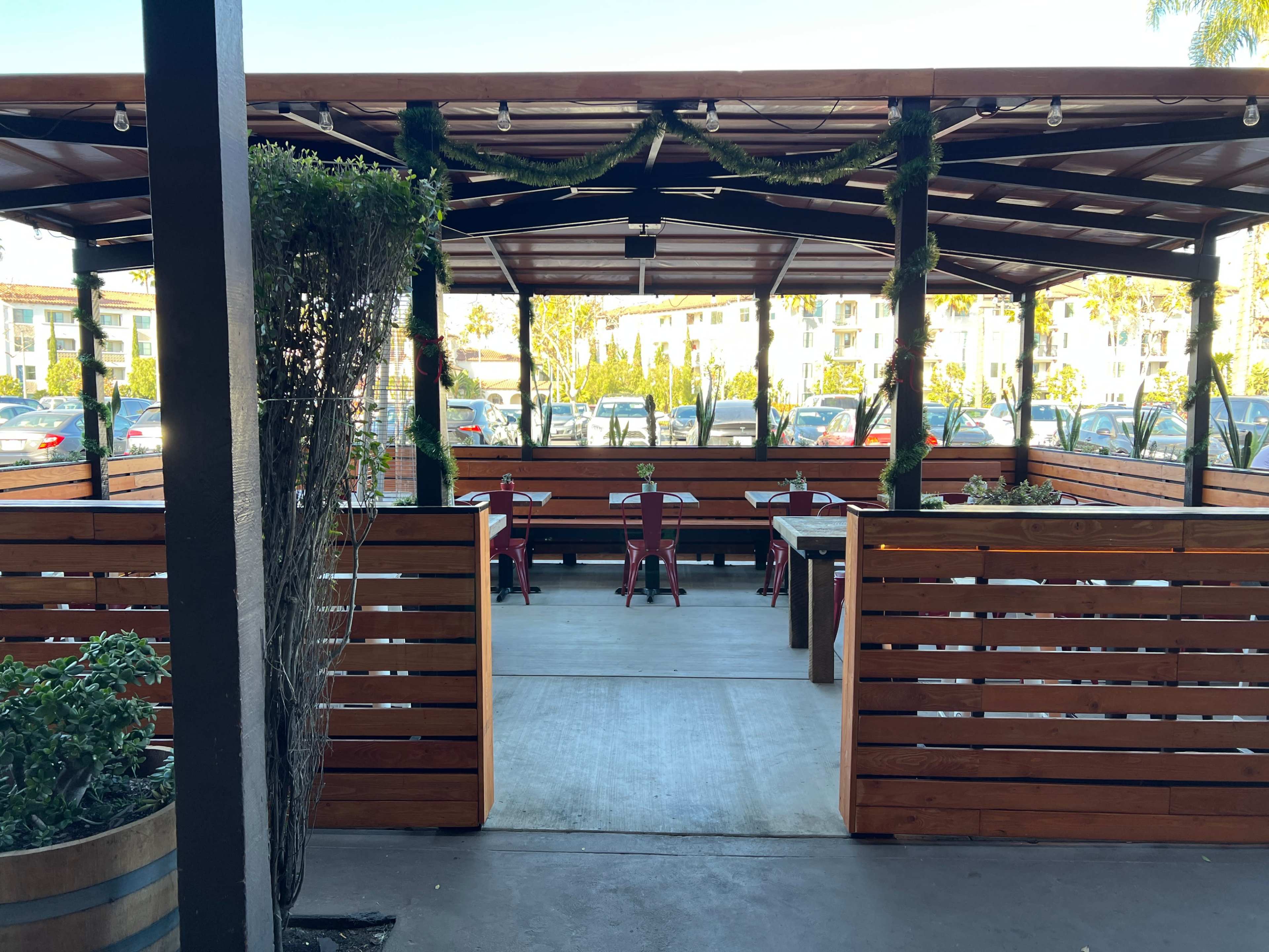 The image shows a covered outdoor dining area with wooden slat walls, tables, and chairs arranged beneath a canopy.