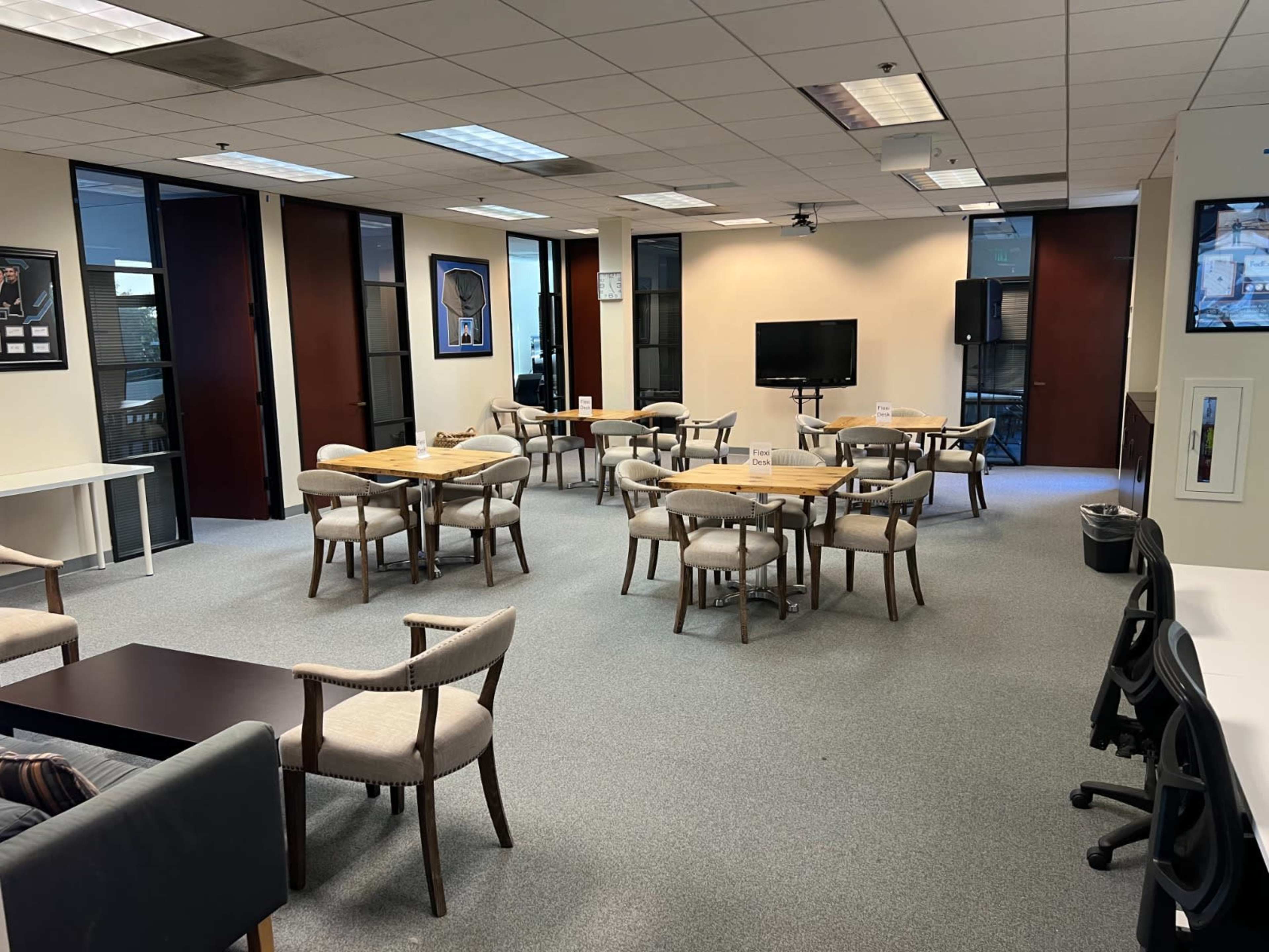 A spacious office area with several tables and chairs, a television on a stand, and large windows letting in natural light.