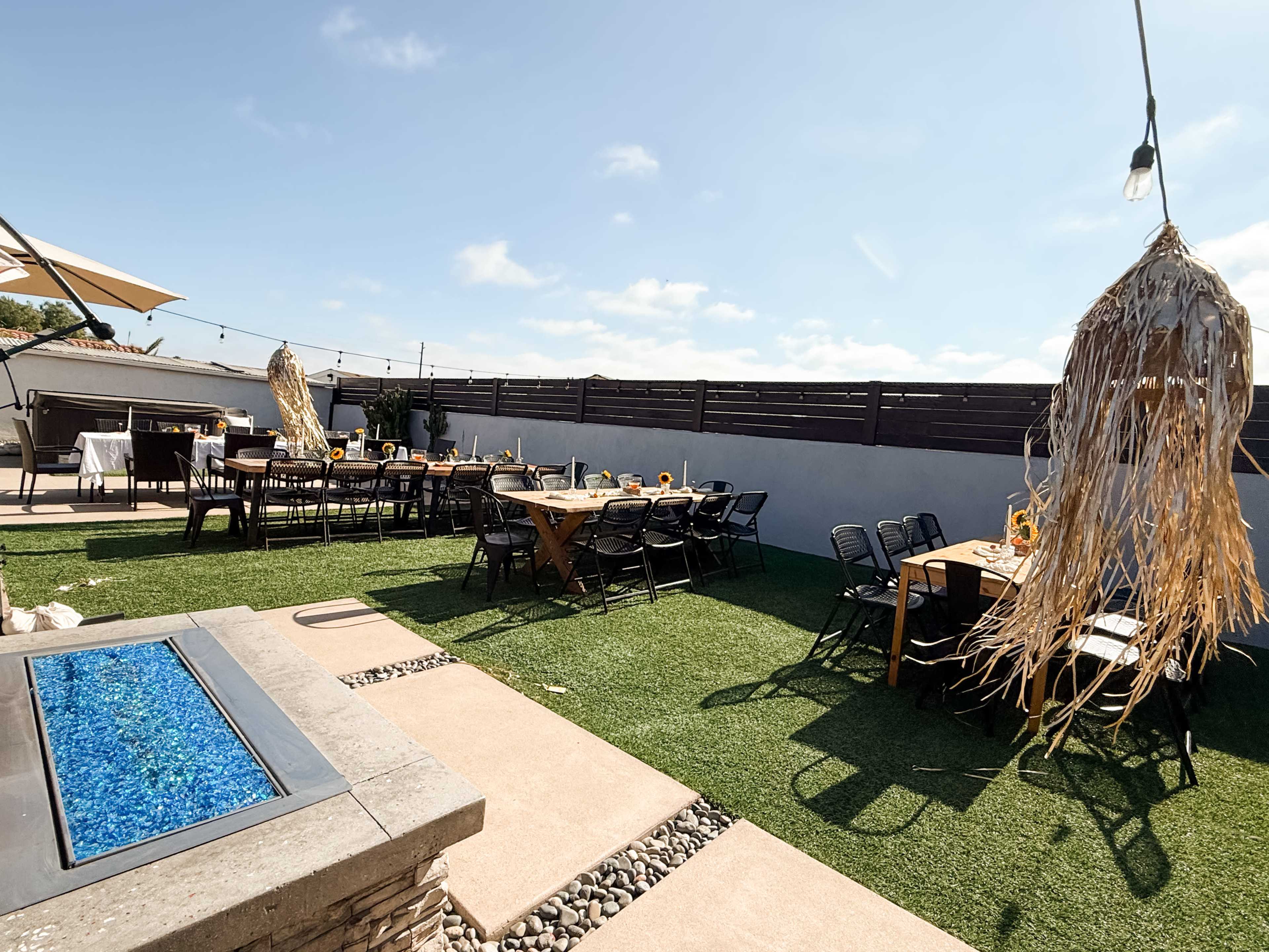 The image shows a spacious outdoor seating area with multiple tables arranged on grassy ground, along with a fire pit and decorative hanging elements.