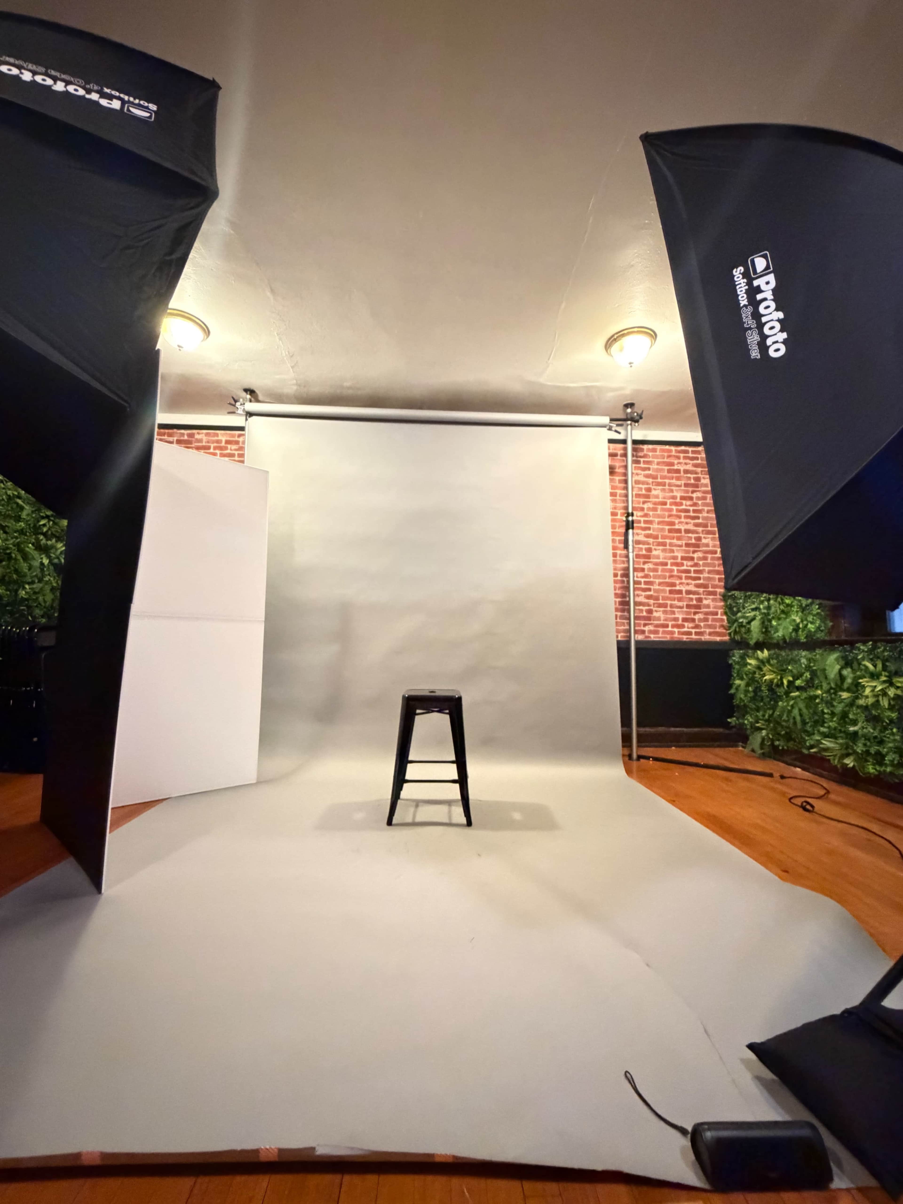The image shows a photography studio setup with a gray backdrop, a black stool in the center, and two softbox lights positioned on either side.