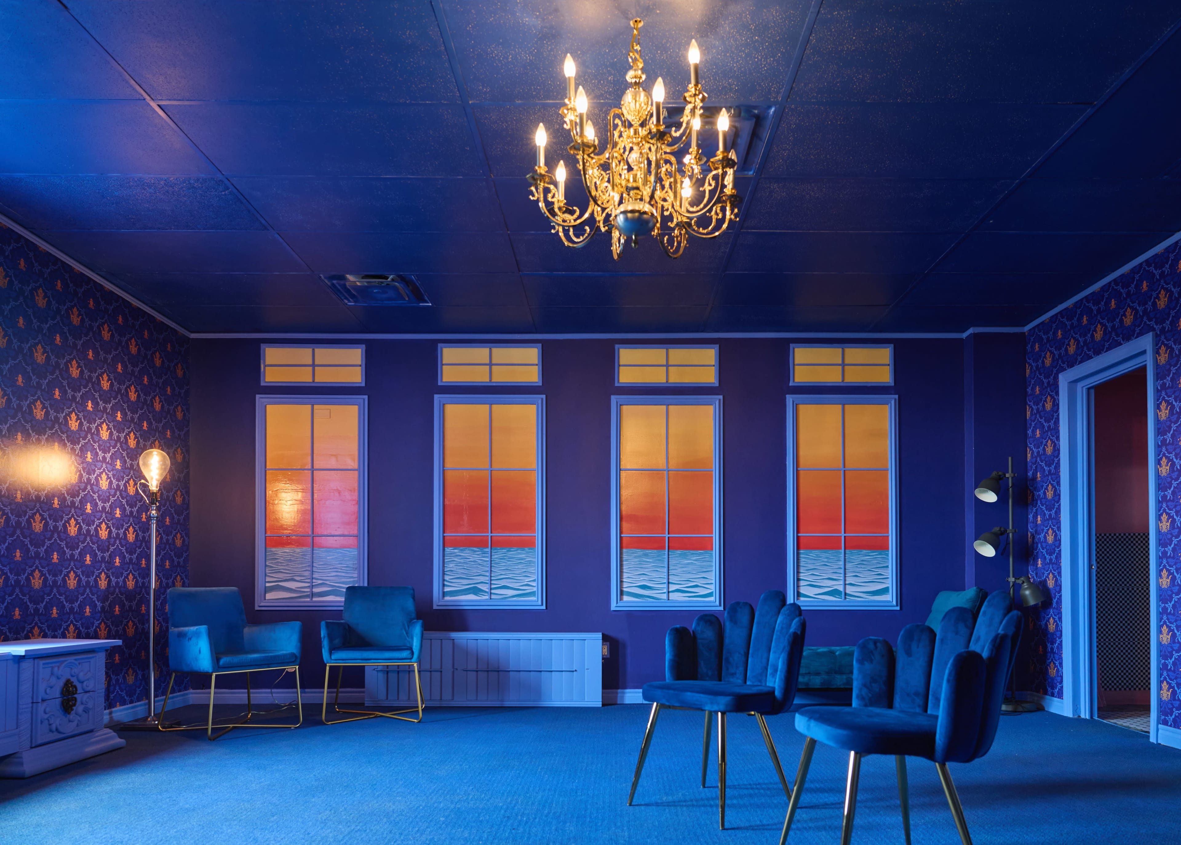 The image shows a room with blue walls adorned with patterned wallpaper, featuring a chandelier and several blue chairs arranged around a carpeted floor.